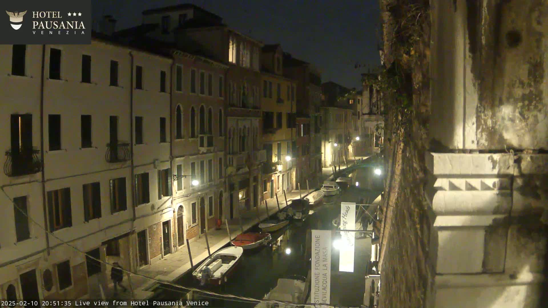 A nighttime view of a narrow canal in Venice, lined with illuminated buildings, with several boats moored along its sides.