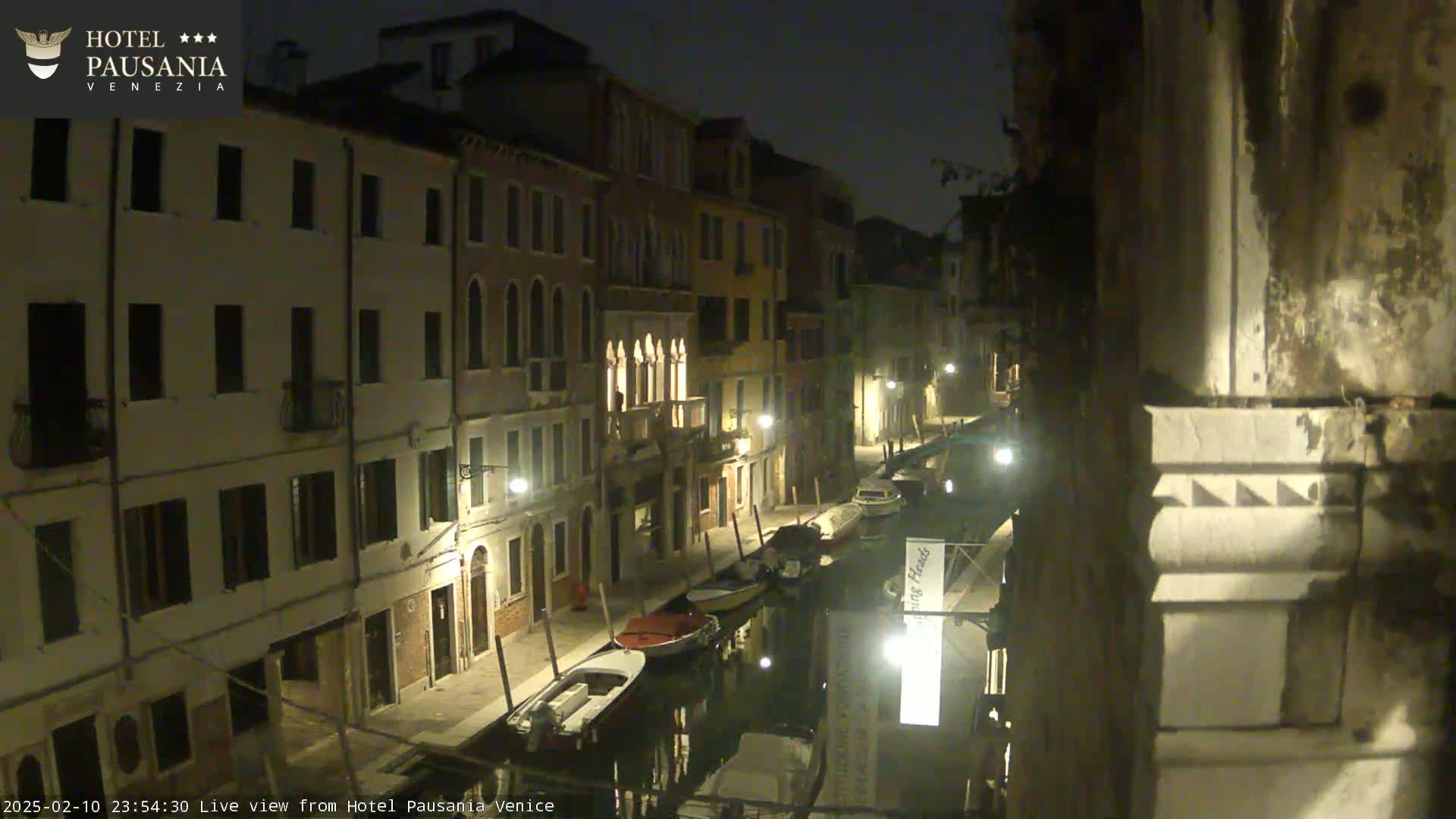 A nighttime view of a narrow canal in Venice, lined with illuminated buildings and several moored boats.