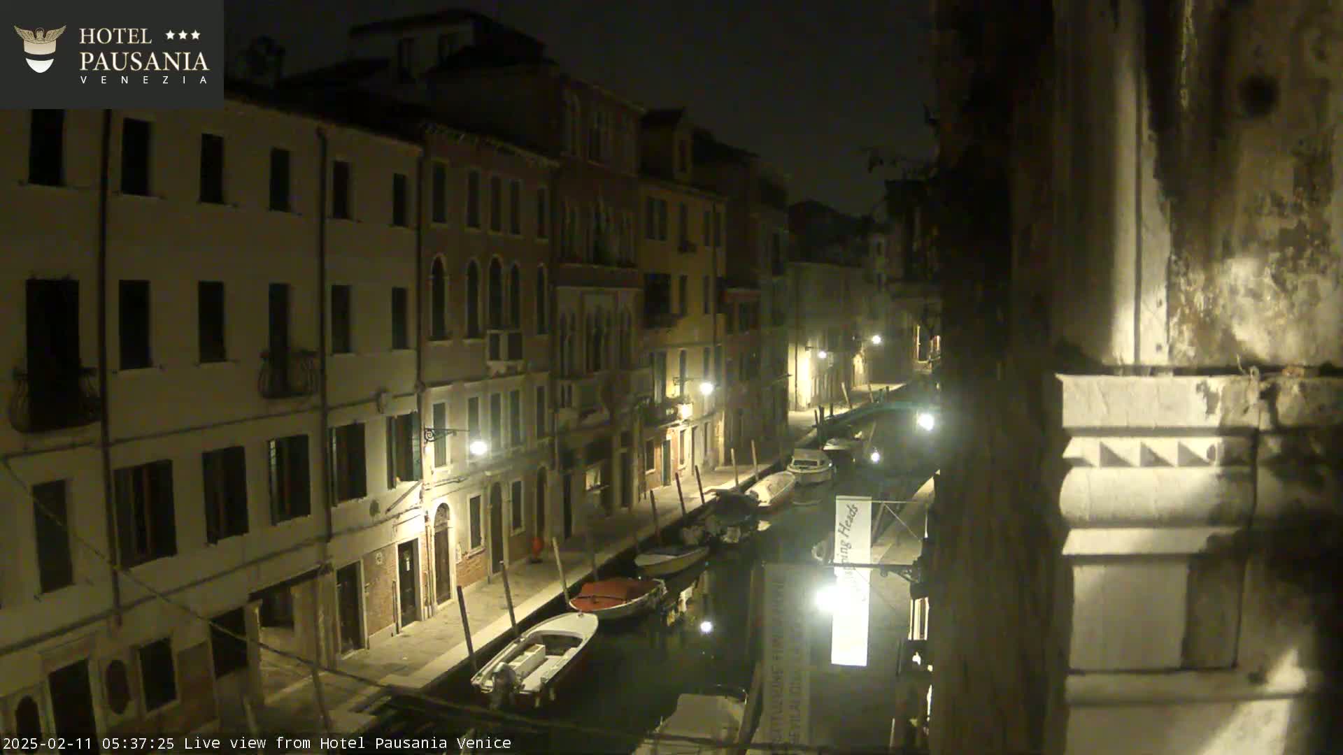 A nighttime view of a narrow canal in Venice, lined with illuminated buildings and several moored boats.