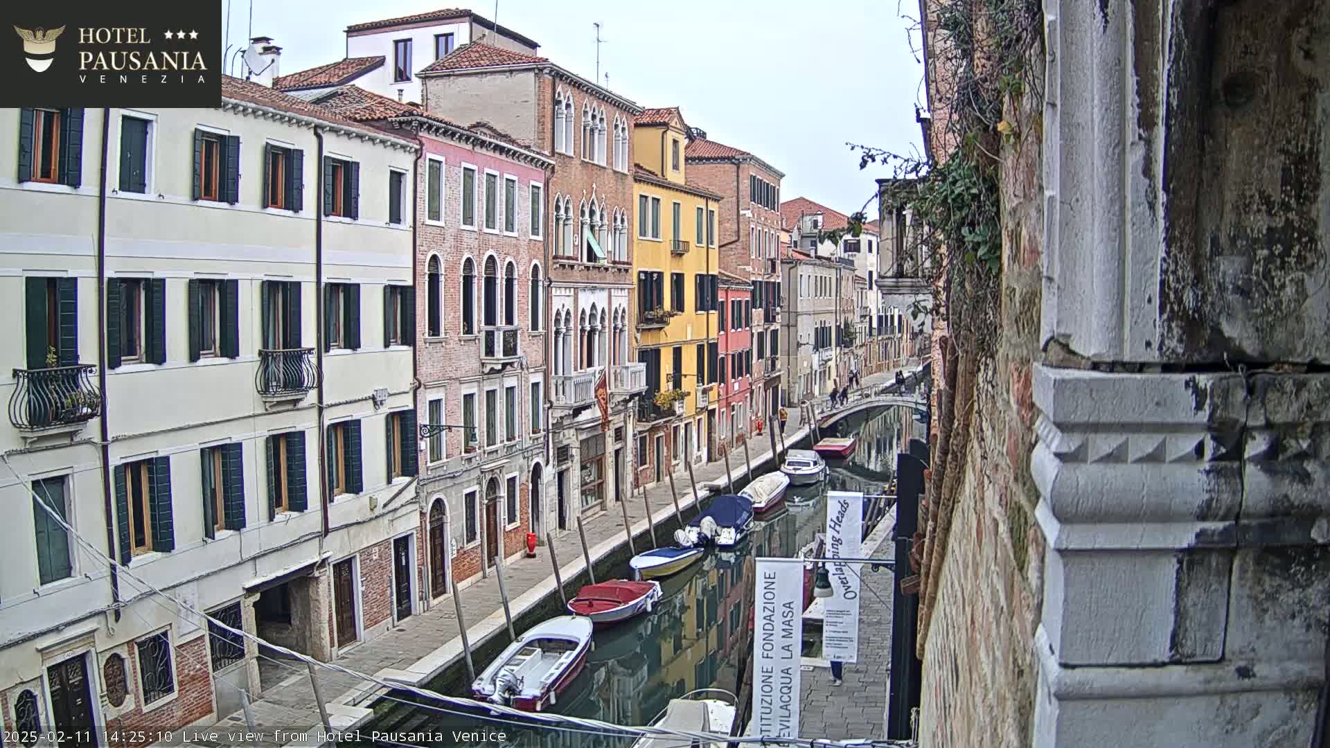 A narrow canal in Venice, Italy, is lined with closely spaced, colorful buildings under an overcast sky; several small boats are moored along the waterway.