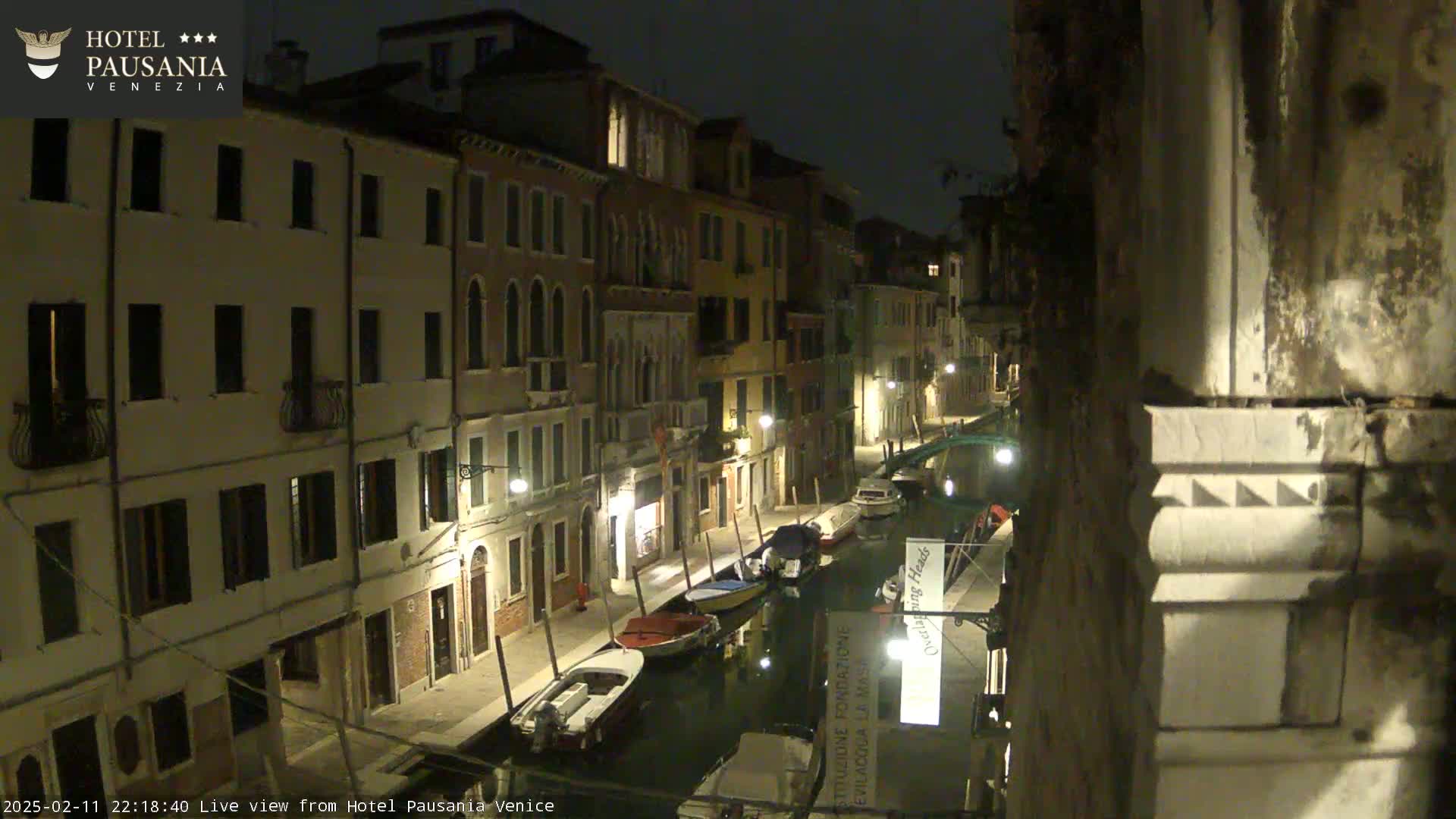 A nighttime view of a narrow canal in Venice, lined with illuminated buildings and several boats moored along the waterway.