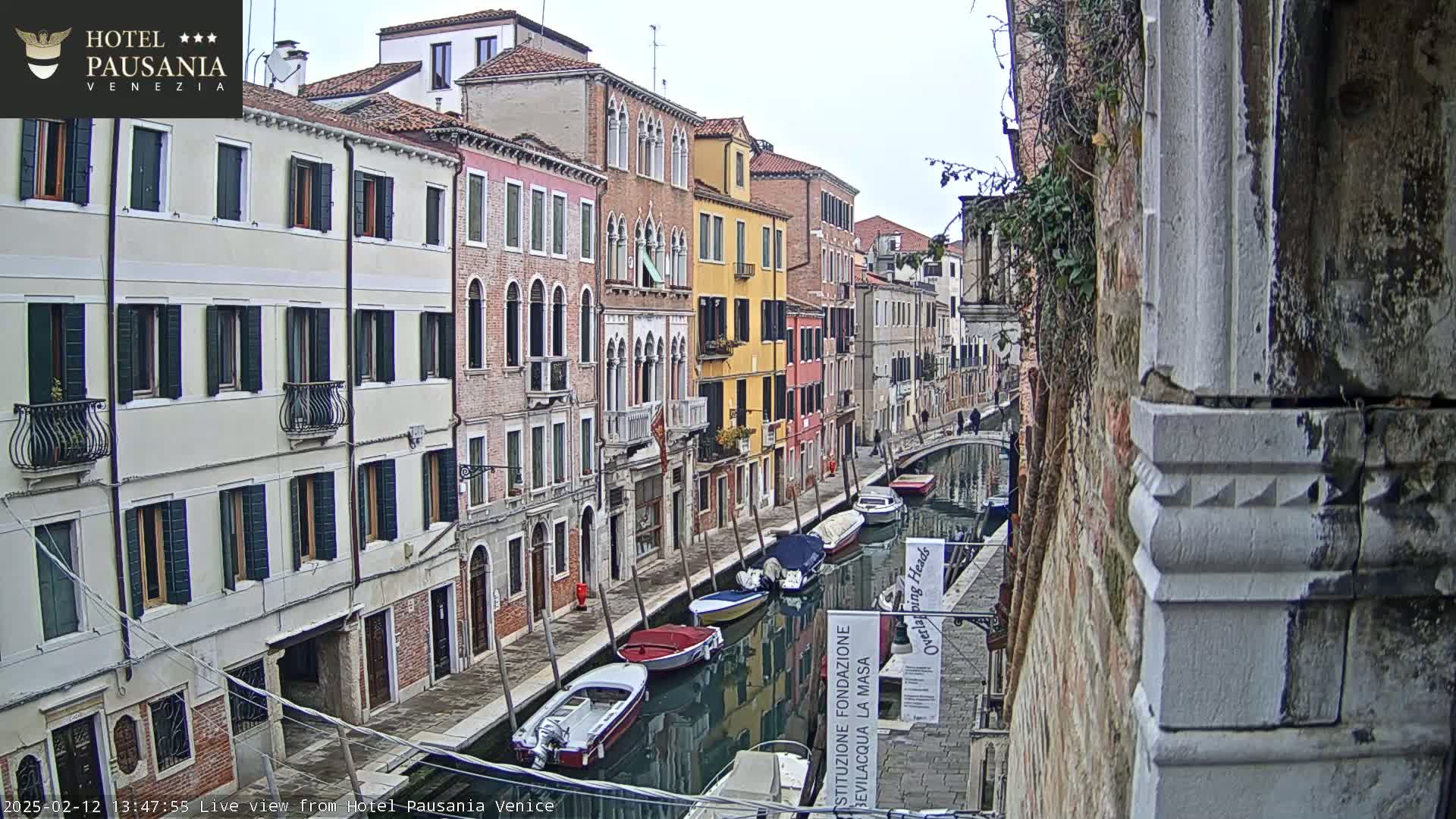 A narrow Venetian canal is lined with colorful buildings under an overcast sky, several small boats are moored along its sides.