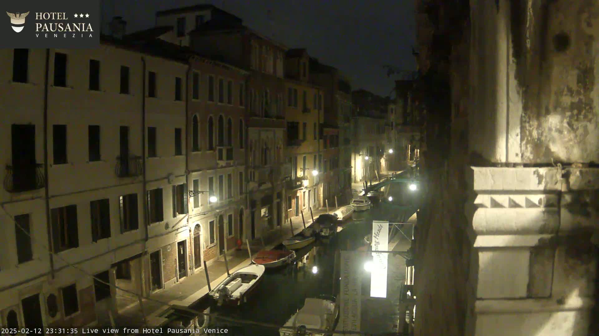 A nighttime view of a narrow canal in Venice, lined with illuminated buildings and moored boats.
