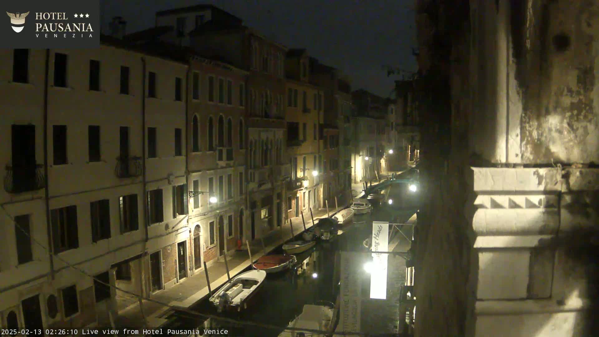 A nighttime view of a narrow canal in Venice, lined with illuminated buildings and several moored boats.