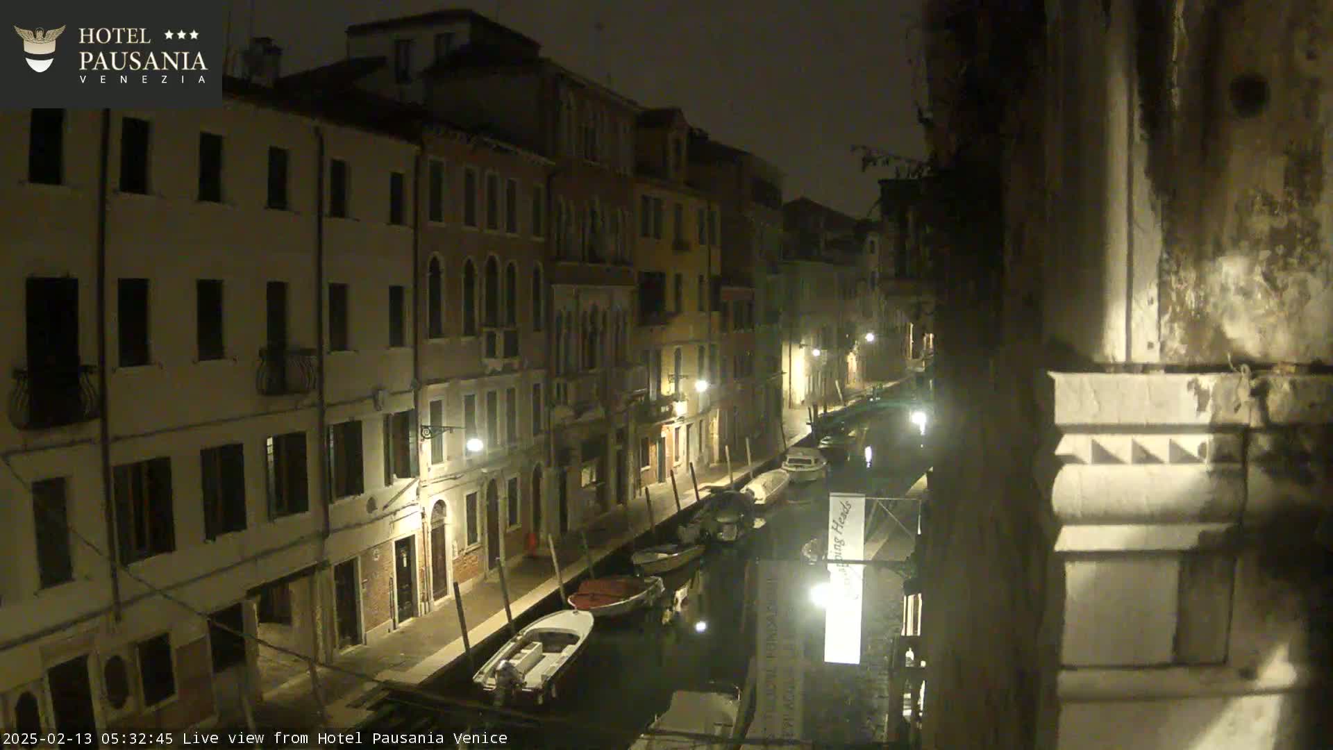 A nighttime view of a narrow canal in Venice, with several boats moored along the sides, and buildings lit by streetlights lining the waterway.