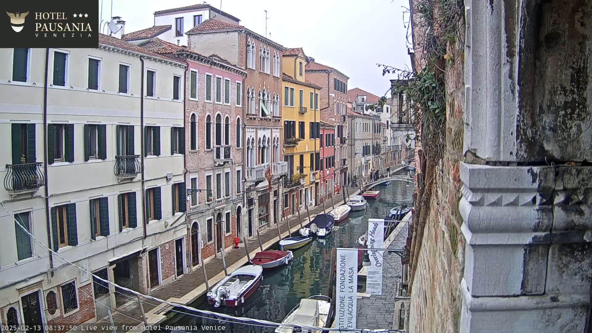 A narrow Venetian canal, lined with multi-story buildings in various colors, shows several boats moored along its sides under an overcast sky.
