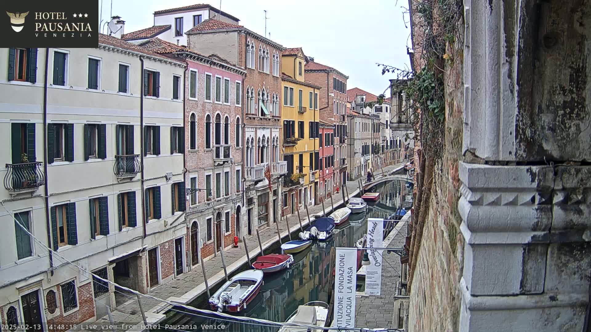 A narrow canal in Venice, Italy, is lined with colorful buildings on a cloudy day, and several small boats are moored along its banks.