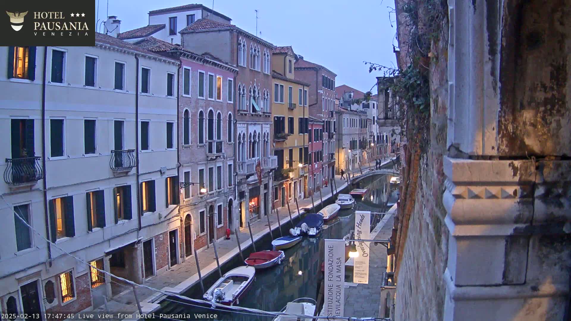 A twilight view of a narrow canal in Venice, lined with closely-spaced buildings, with several boats moored along its sides.