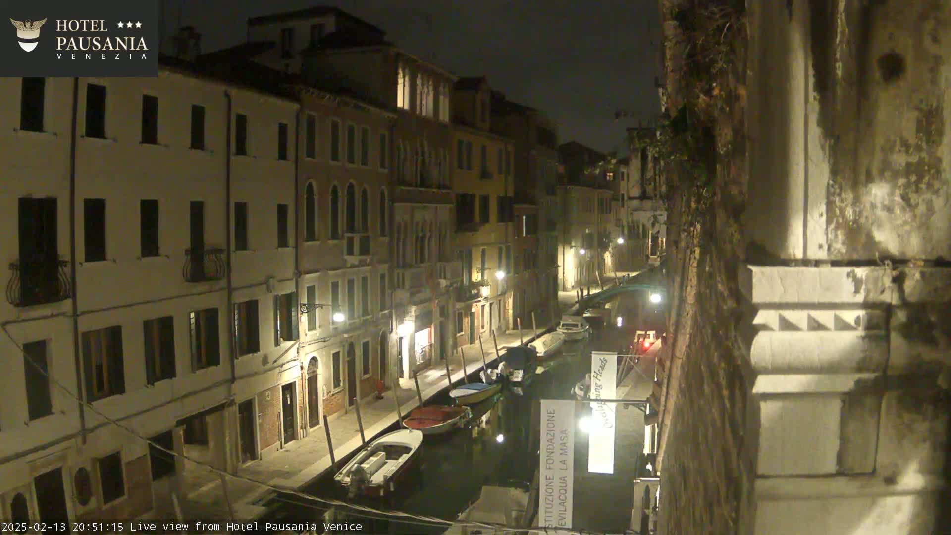 A nighttime view of a narrow canal in Venice, lined with buildings and several moored boats.