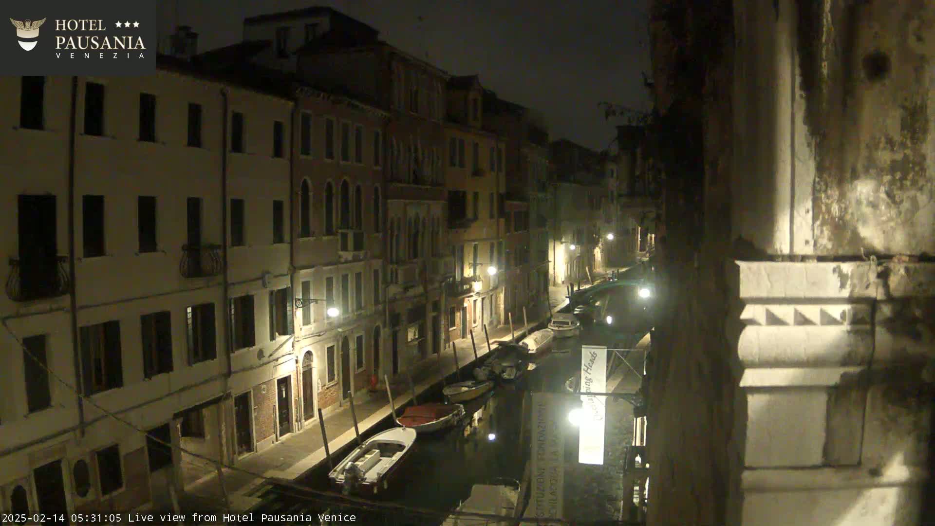 A nighttime view of a narrow canal in Venice, lined with buildings, with several boats moored along its sides.