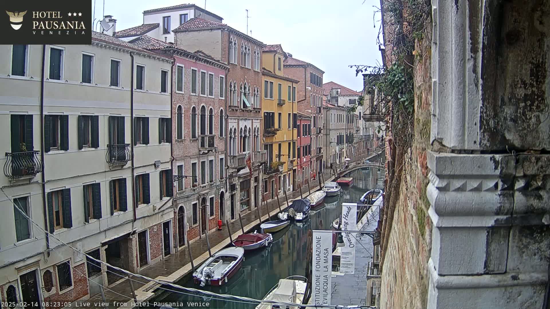 A narrow canal in Venice, Italy, shows several moored boats under an overcast sky and light rain, flanked by tall, colorful buildings.