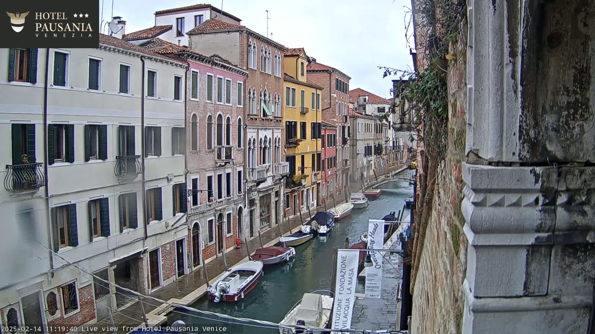 A canal in Venice, Italy, with several boats moored along its sides, is viewed on an overcast day from a high vantage point showing colorful buildings lining the waterway.