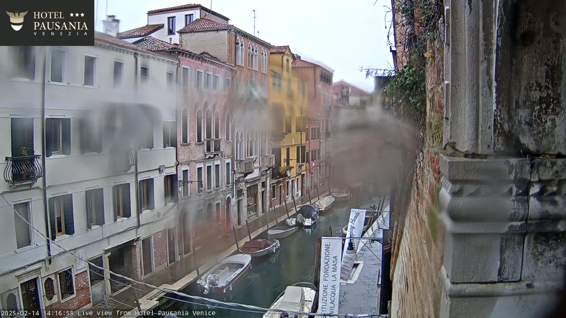 A view of a canal in Venice, Italy, on a misty or foggy day, with several boats moored along the sides of the canal and buildings lining the waterway.
