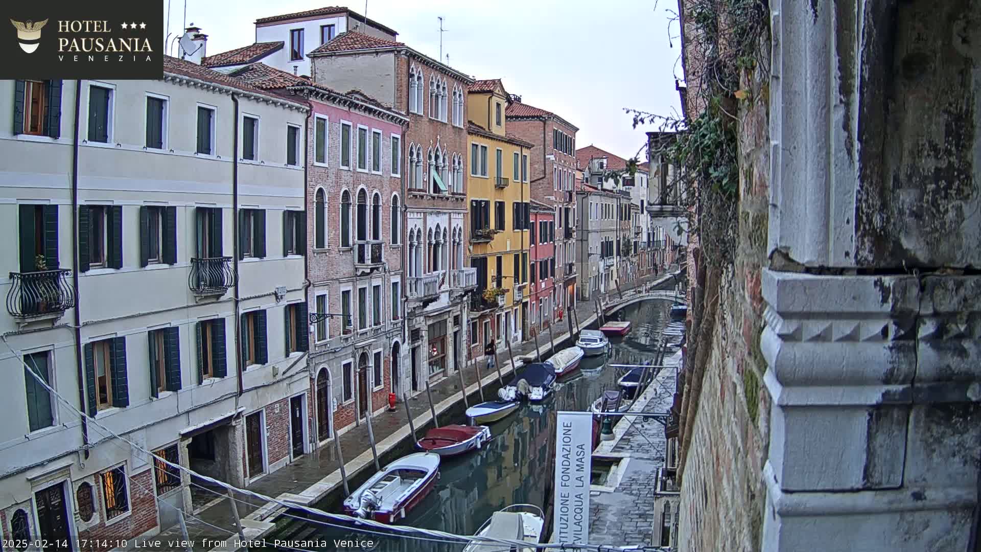 A narrow canal in Venice, Italy, is lined with colorful buildings under an overcast sky, and several boats are moored along the waterway.