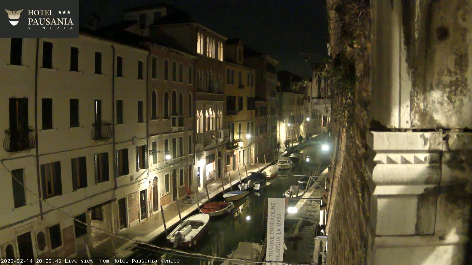A nighttime view of a narrow canal in Venice, lined with illuminated buildings and several moored boats.