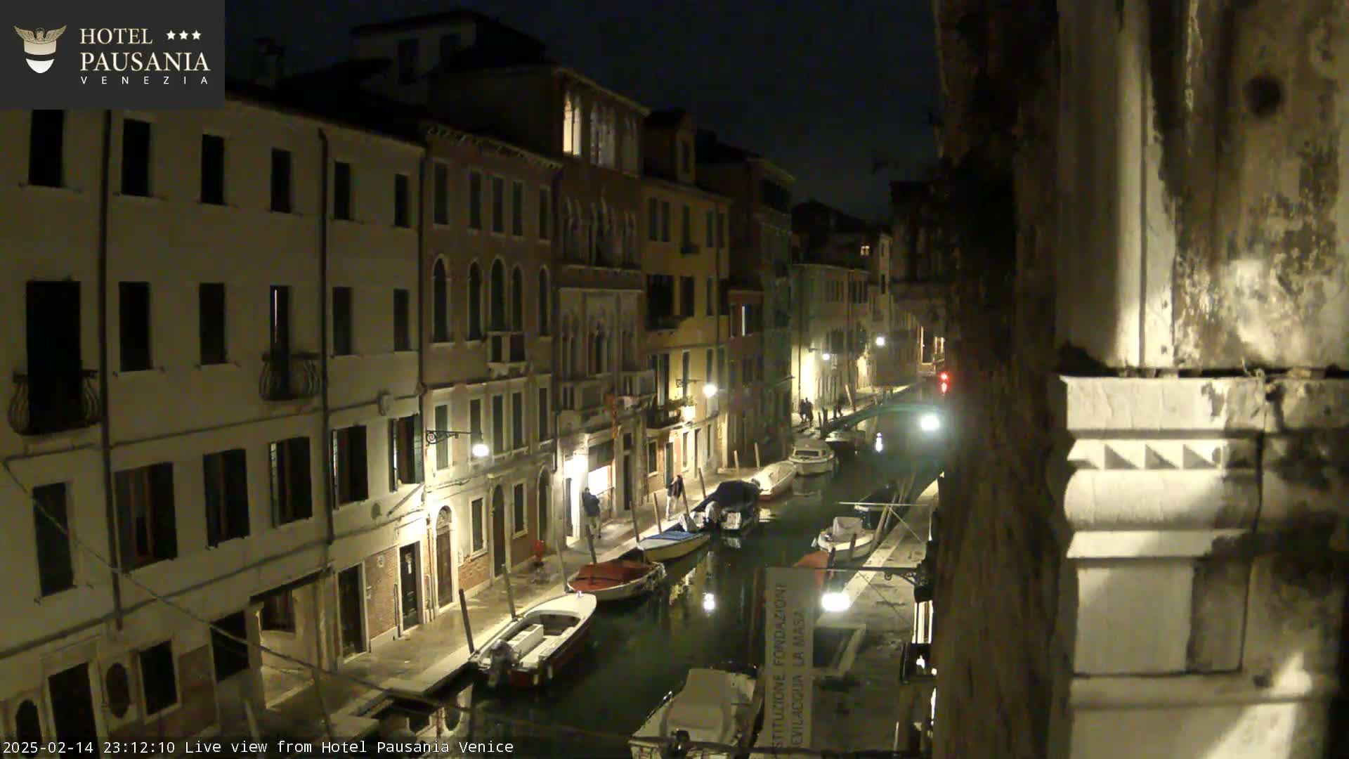 A nighttime view of a narrow canal in Venice, lined with illuminated buildings and several small boats moored along its banks.