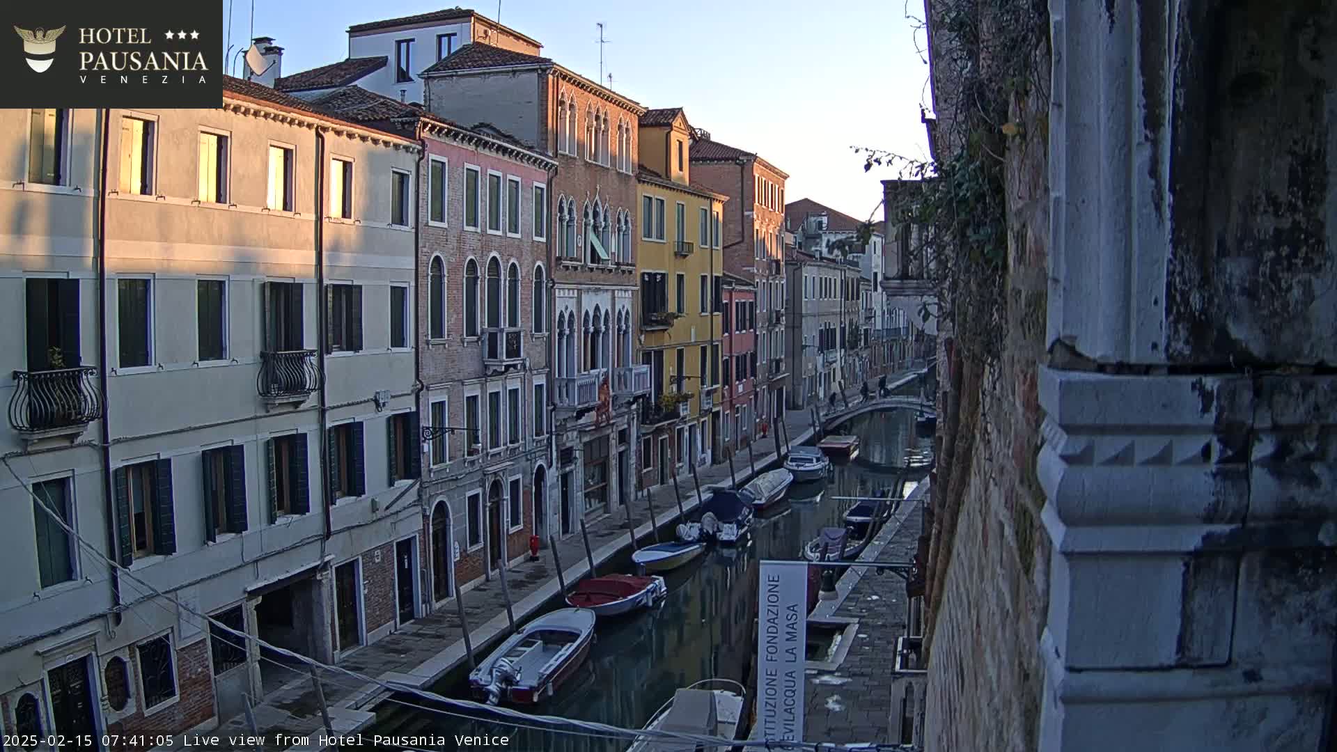 Several boats are moored along a narrow canal lined with tall, closely-spaced buildings at sunrise.