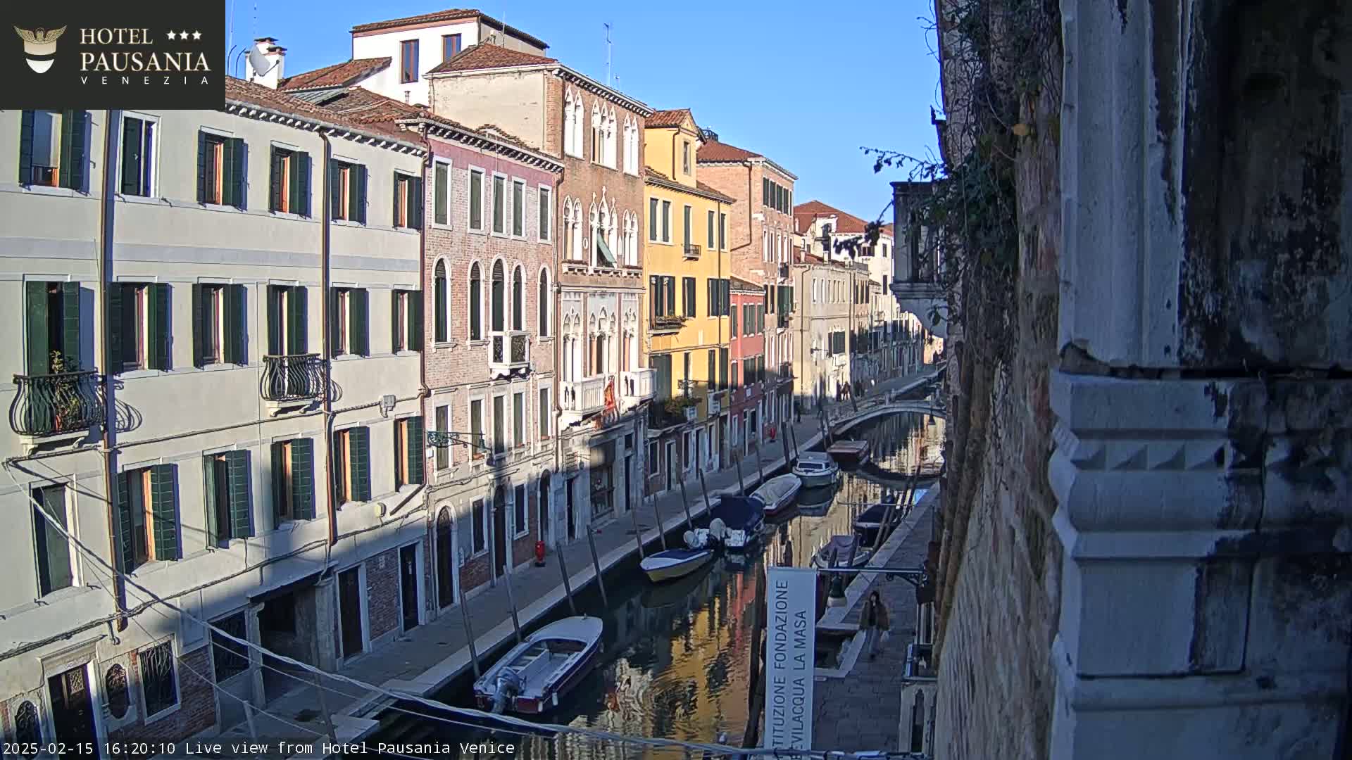 A sunny day in Venice shows several boats moored along a canal beside colorful buildings.