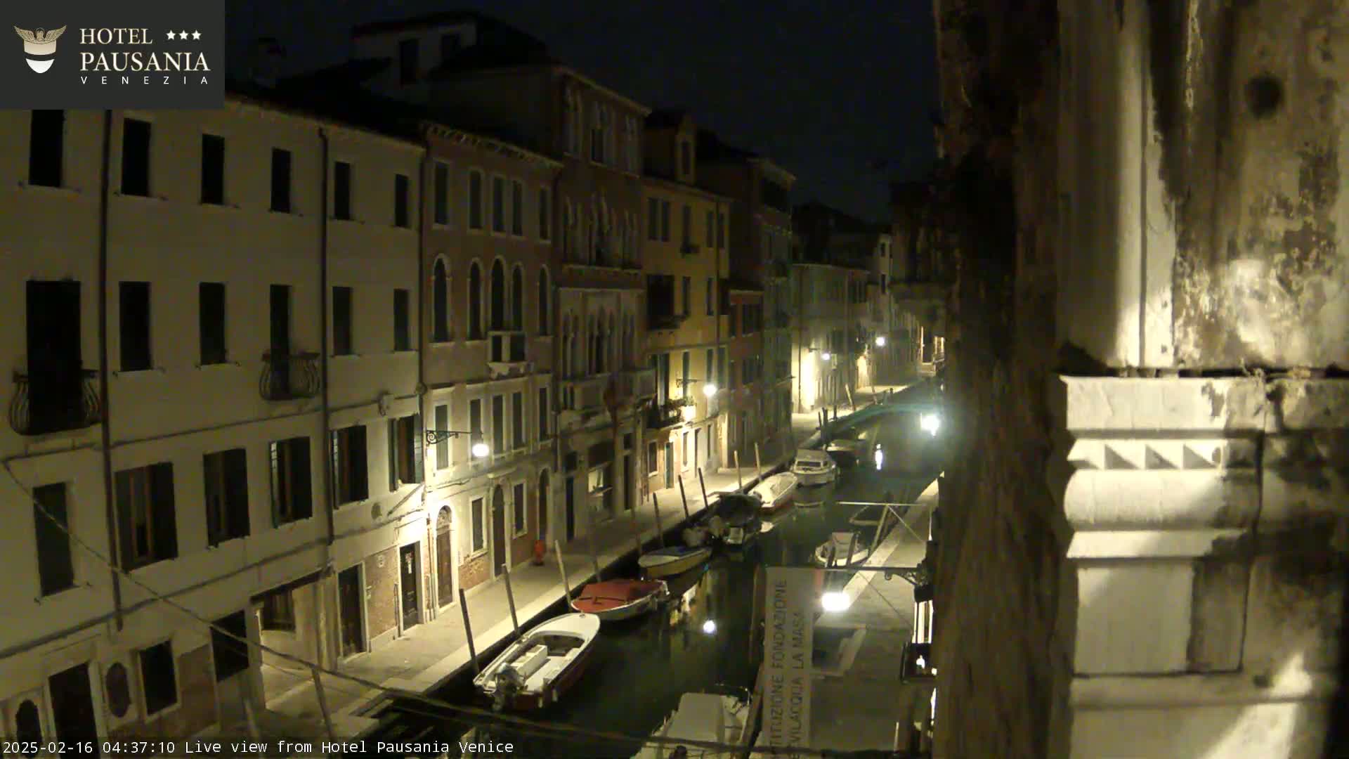 A nighttime view of a narrow canal in Venice, Italy, lined with buildings and several moored boats under a dark sky.