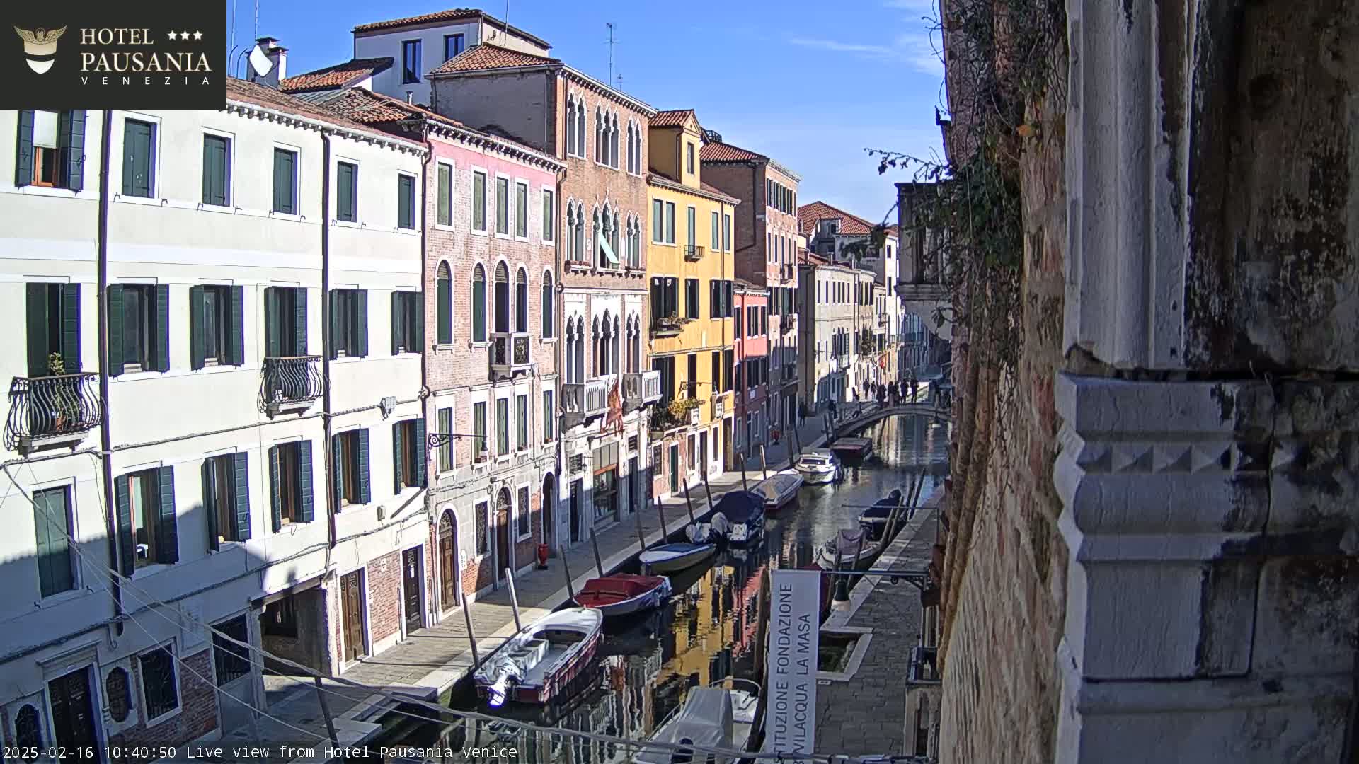 A sunny view of a narrow canal in Venice, Italy, lined with colorful buildings and several boats moored along its edge.