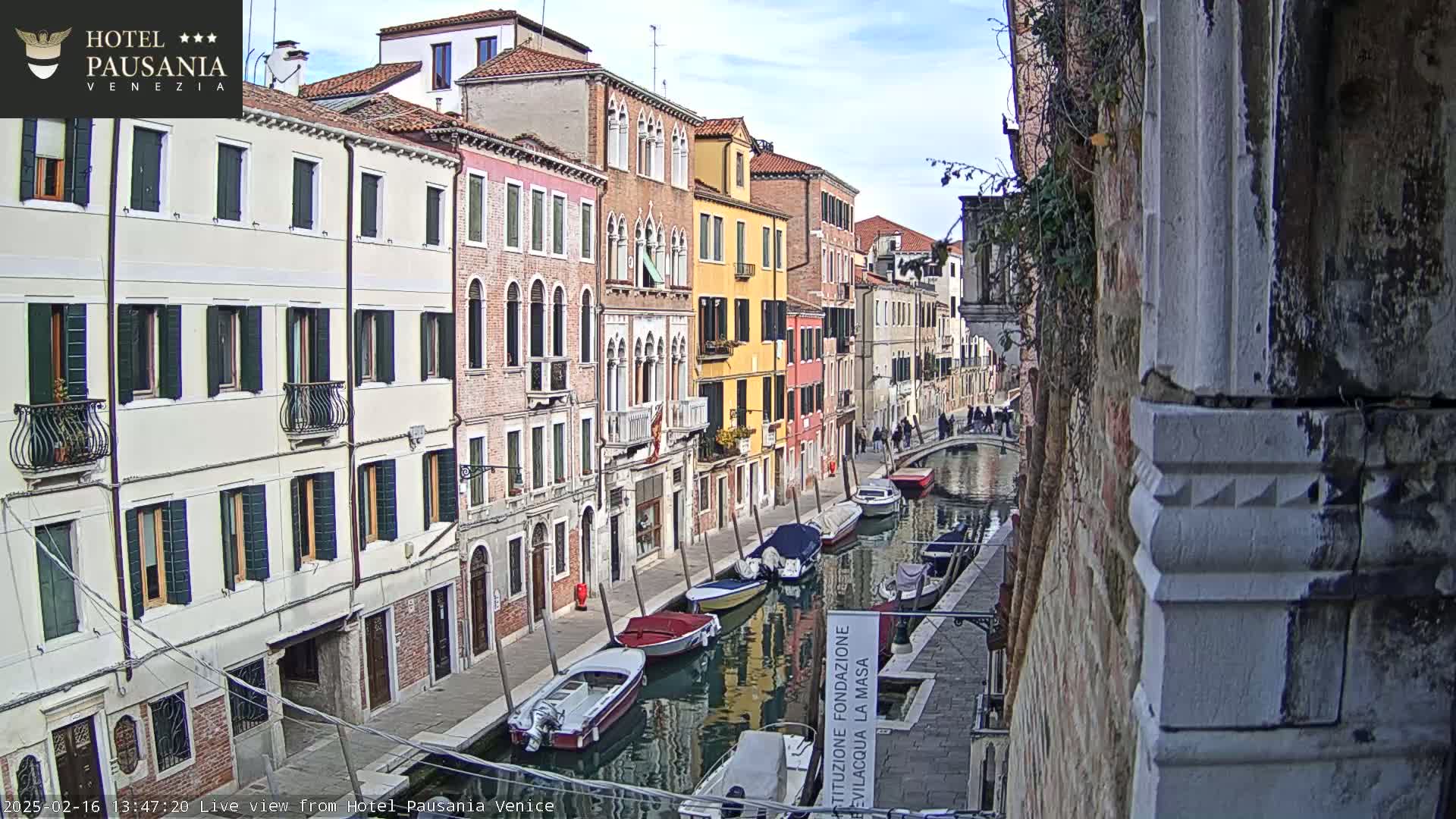 A narrow Venetian canal is lined with colorful buildings under a partly cloudy sky, with several boats moored along its sides.