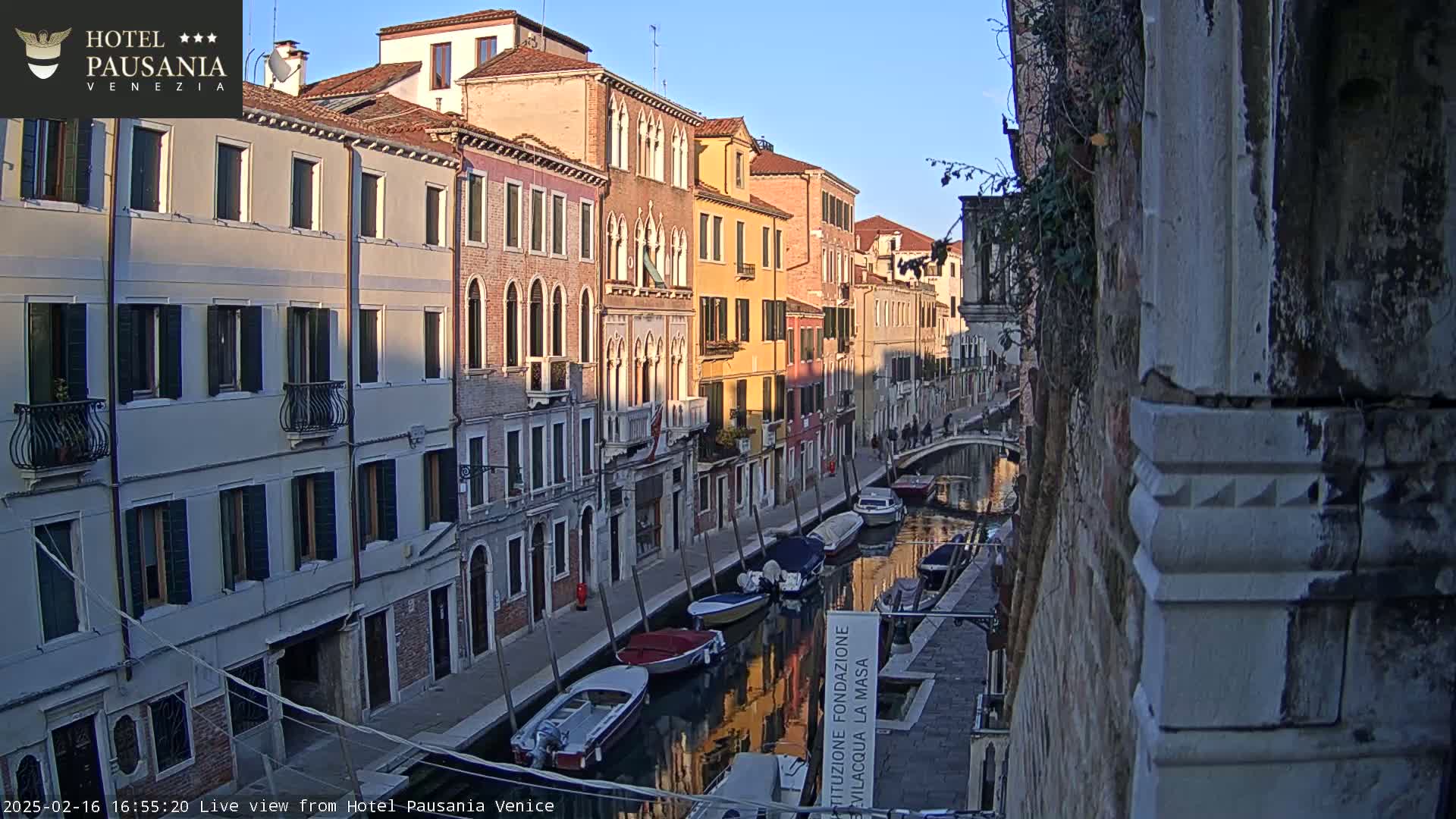 A narrow canal in Venice, Italy, is lined with tall, closely spaced buildings reflecting in the calm water under a clear, bright sky.