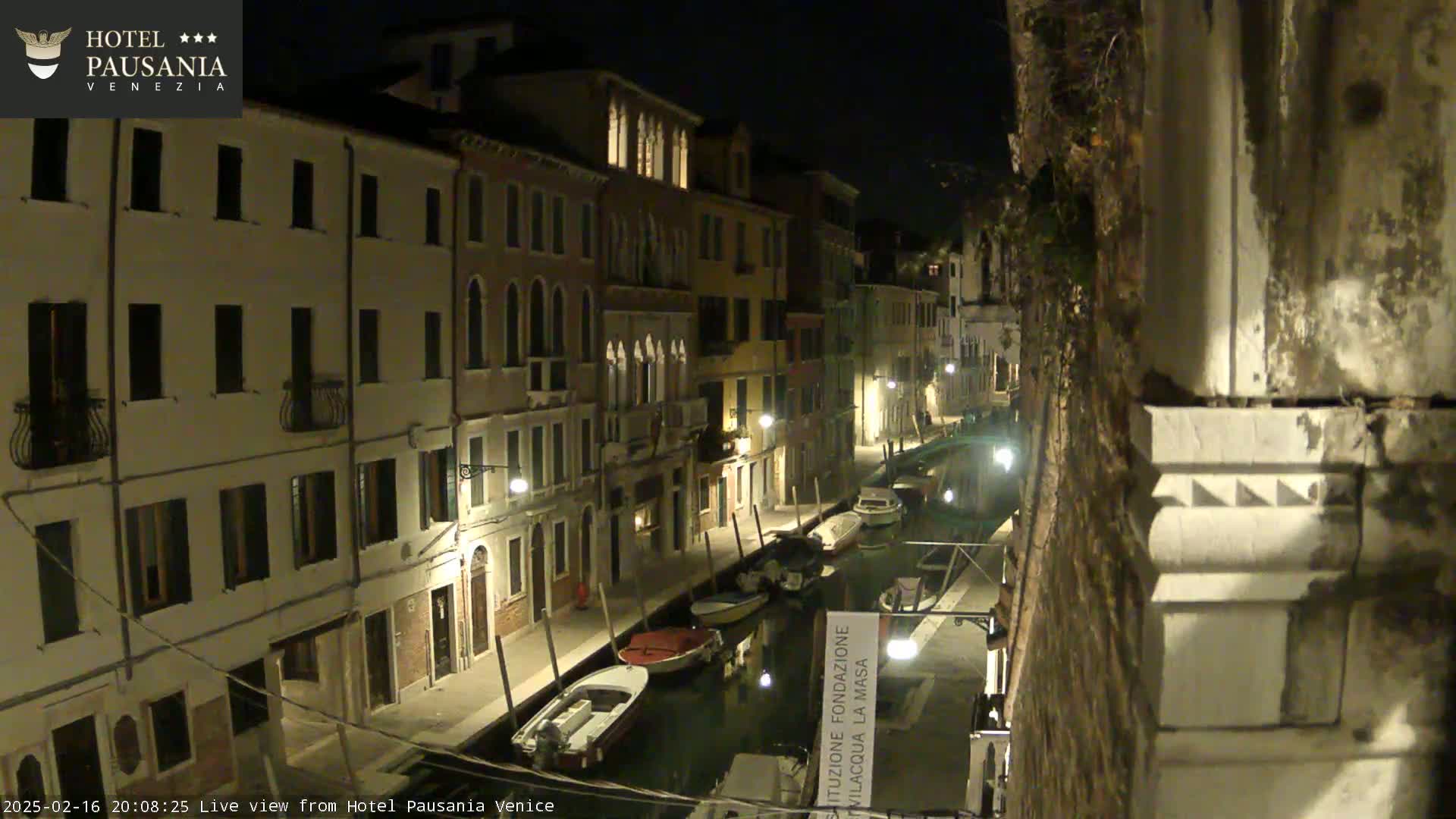 A nighttime view of a narrow canal in Venice, lined with illuminated buildings and moored boats.