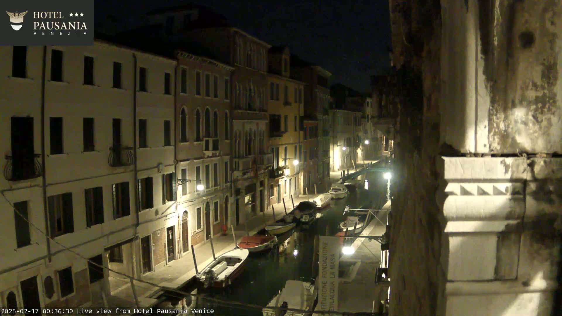 A nighttime view of a canal in Venice, Italy, lined with illuminated buildings and several moored boats.