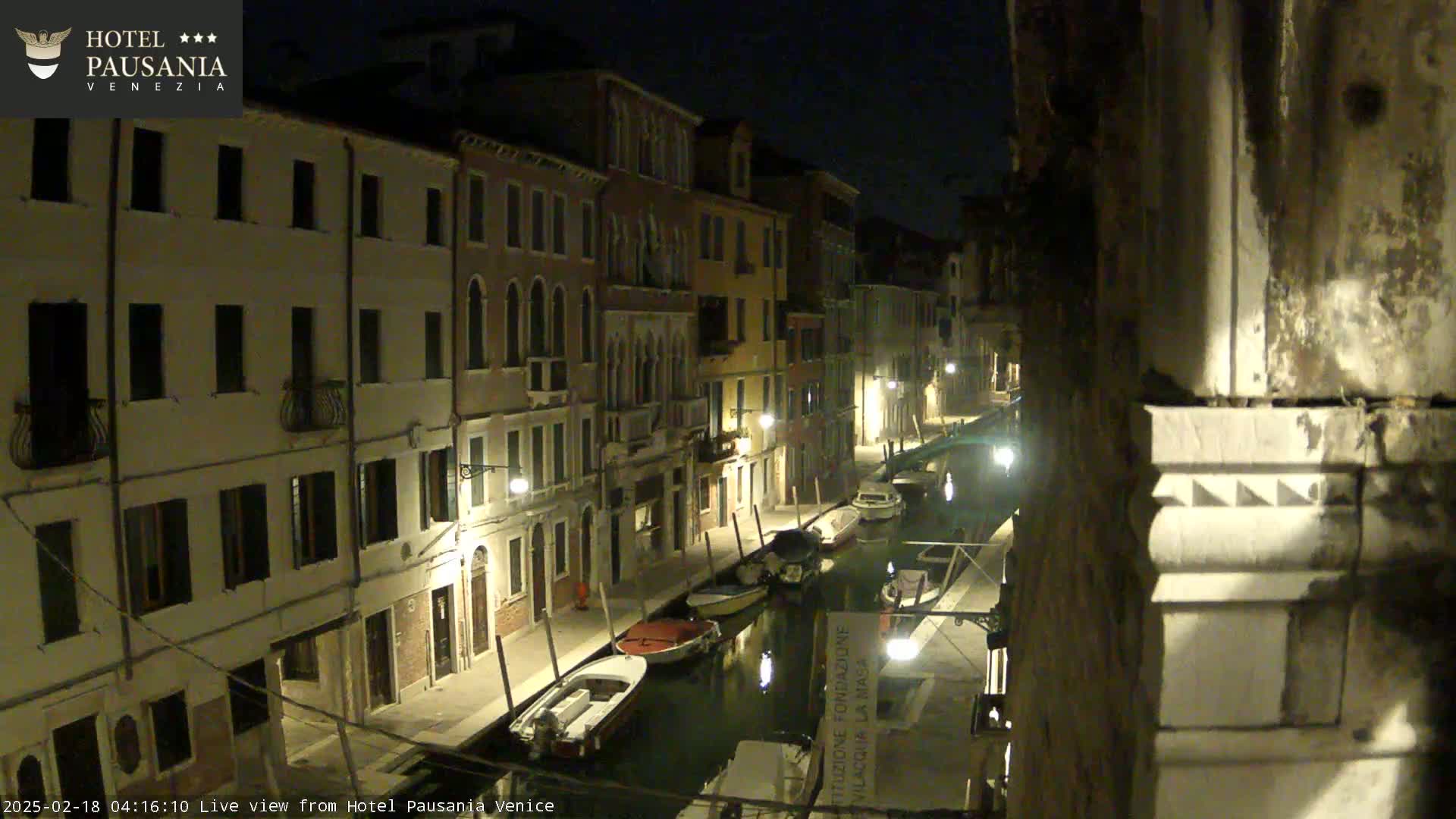 A nighttime view of a narrow canal in Venice, lined with illuminated buildings and several moored boats.