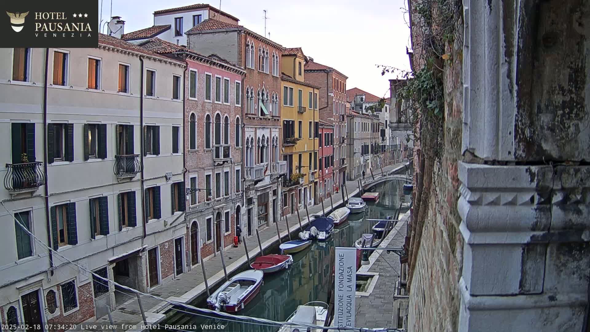 A calm canal in Venice, lined with colorful buildings, is filled with several moored boats under an overcast sky.