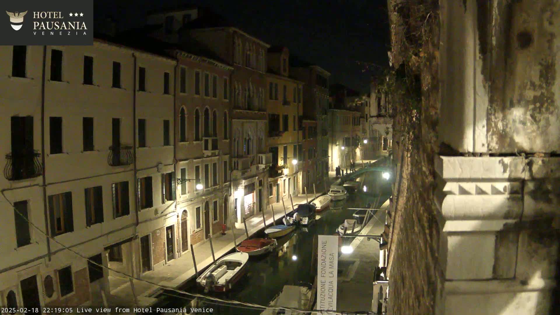 A nighttime view of a narrow canal in Venice, lined with illuminated buildings and several moored boats.