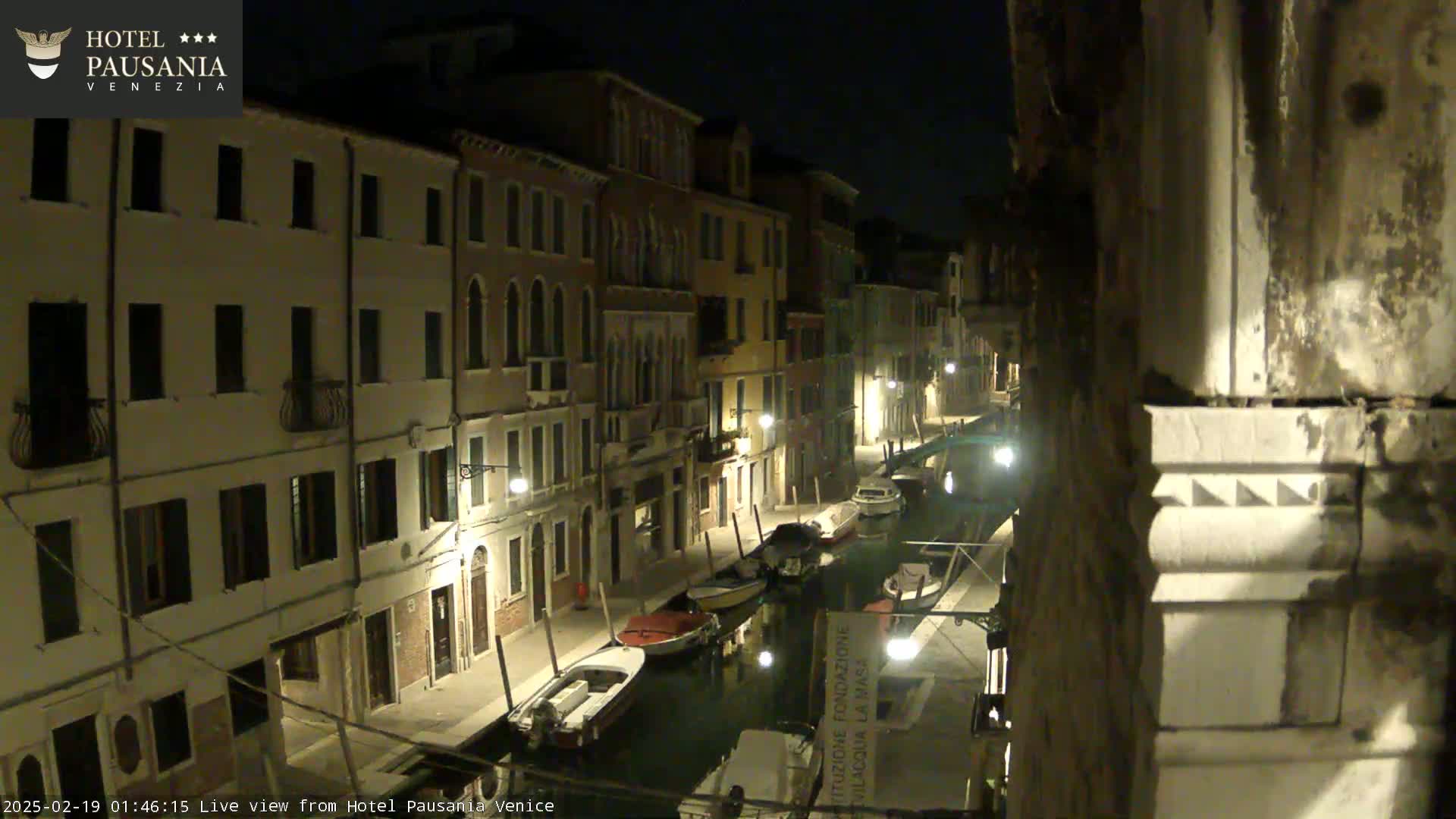 A nighttime view of a narrow canal in Venice, Italy, lined with buildings and several moored boats.