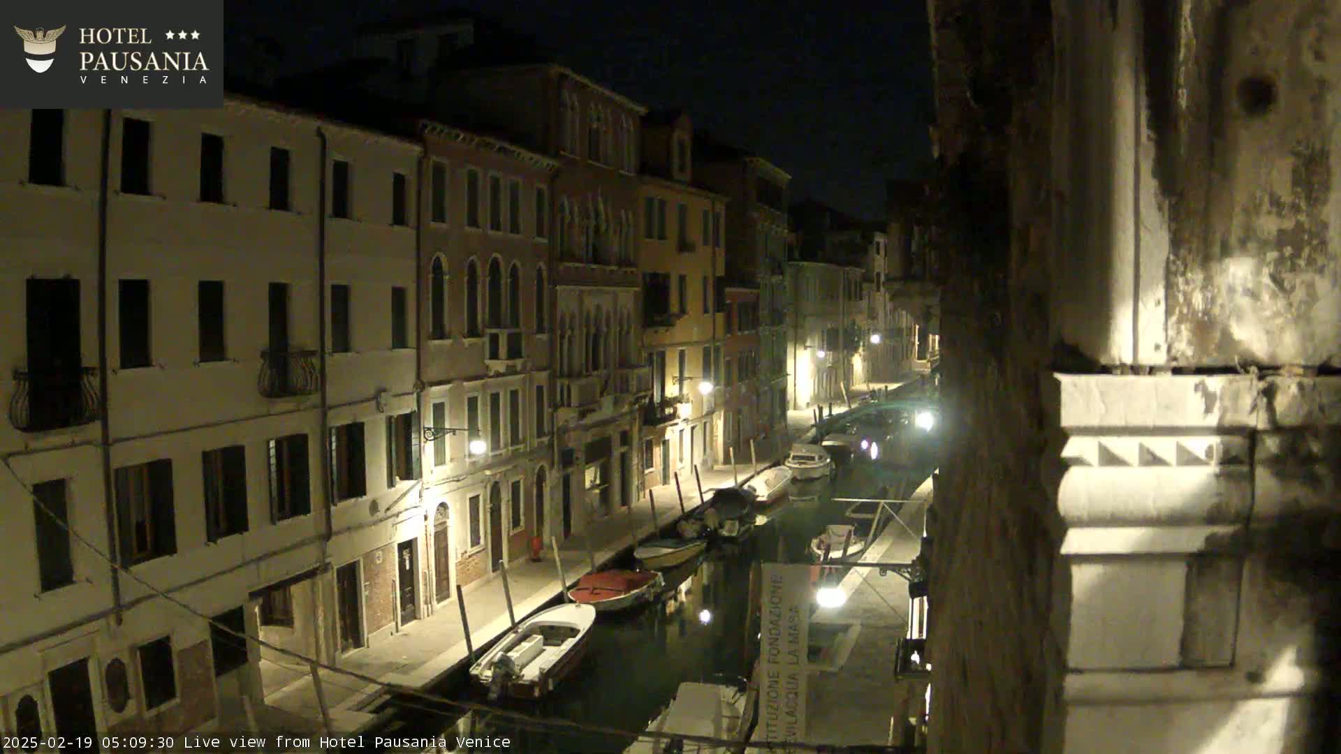 A nighttime view of a canal in Venice, Italy, with several boats moored along its banks, lined by illuminated buildings.