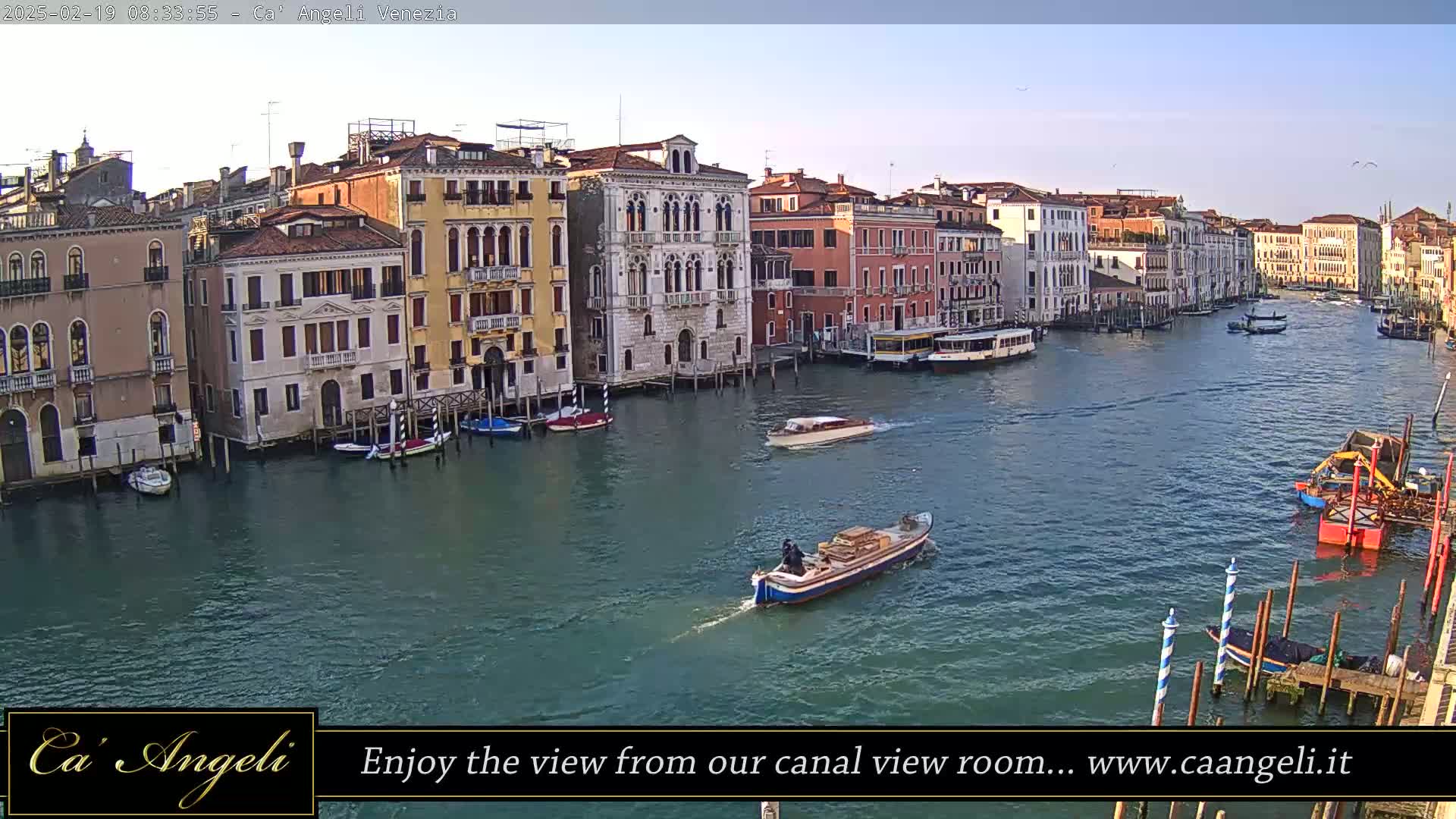 A wide shot of a calm canal in Venice, Italy, shows several buildings lining the waterway, with boats and gondolas traveling on the water under a clear sky.