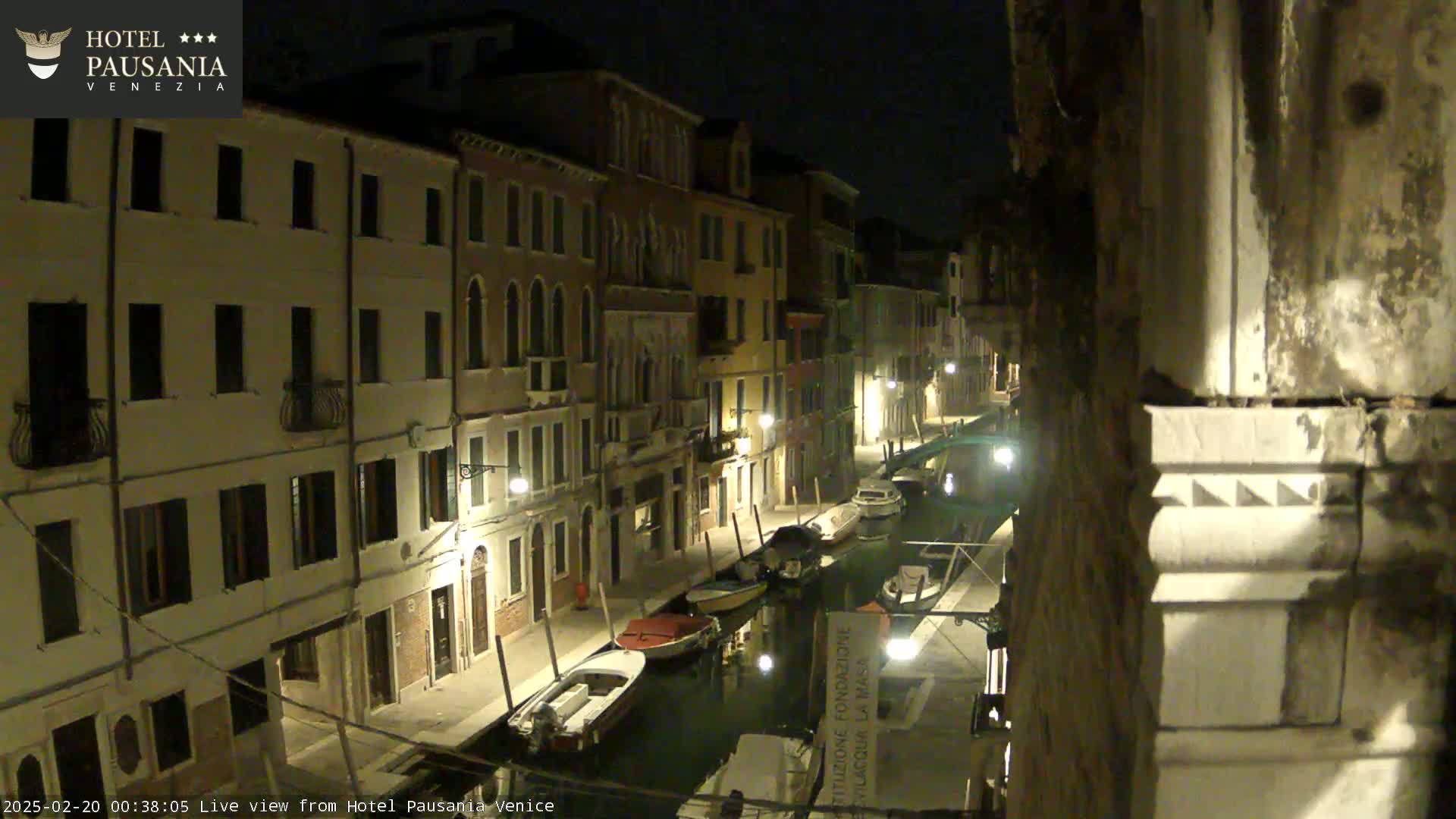 A nighttime view of a canal in Venice, with several boats moored along its banks, lined by illuminated buildings.