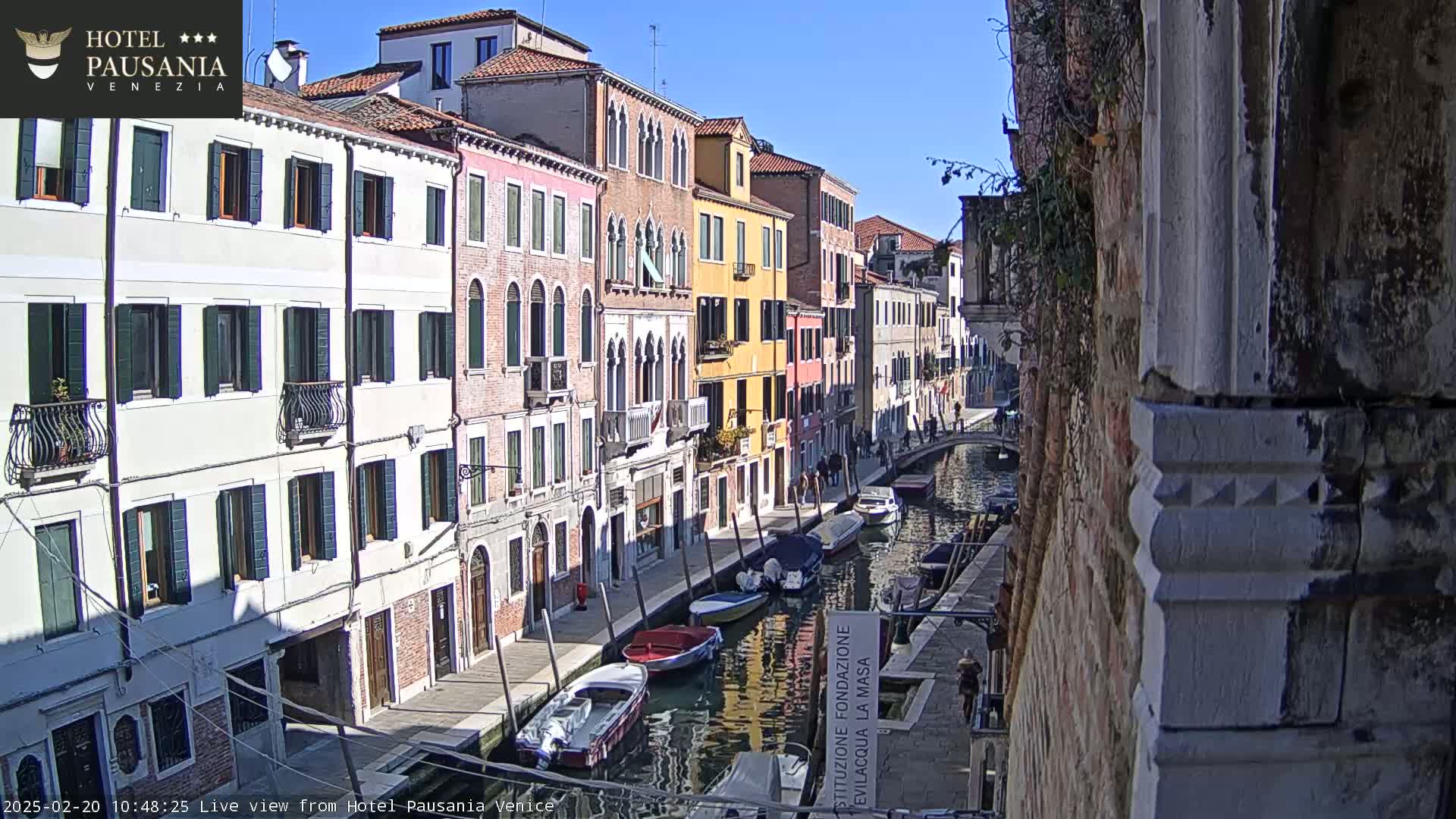 A sunny day in Venice shows a narrow canal lined with colorful buildings, several boats moored along its sides, and people walking along the waterfront.