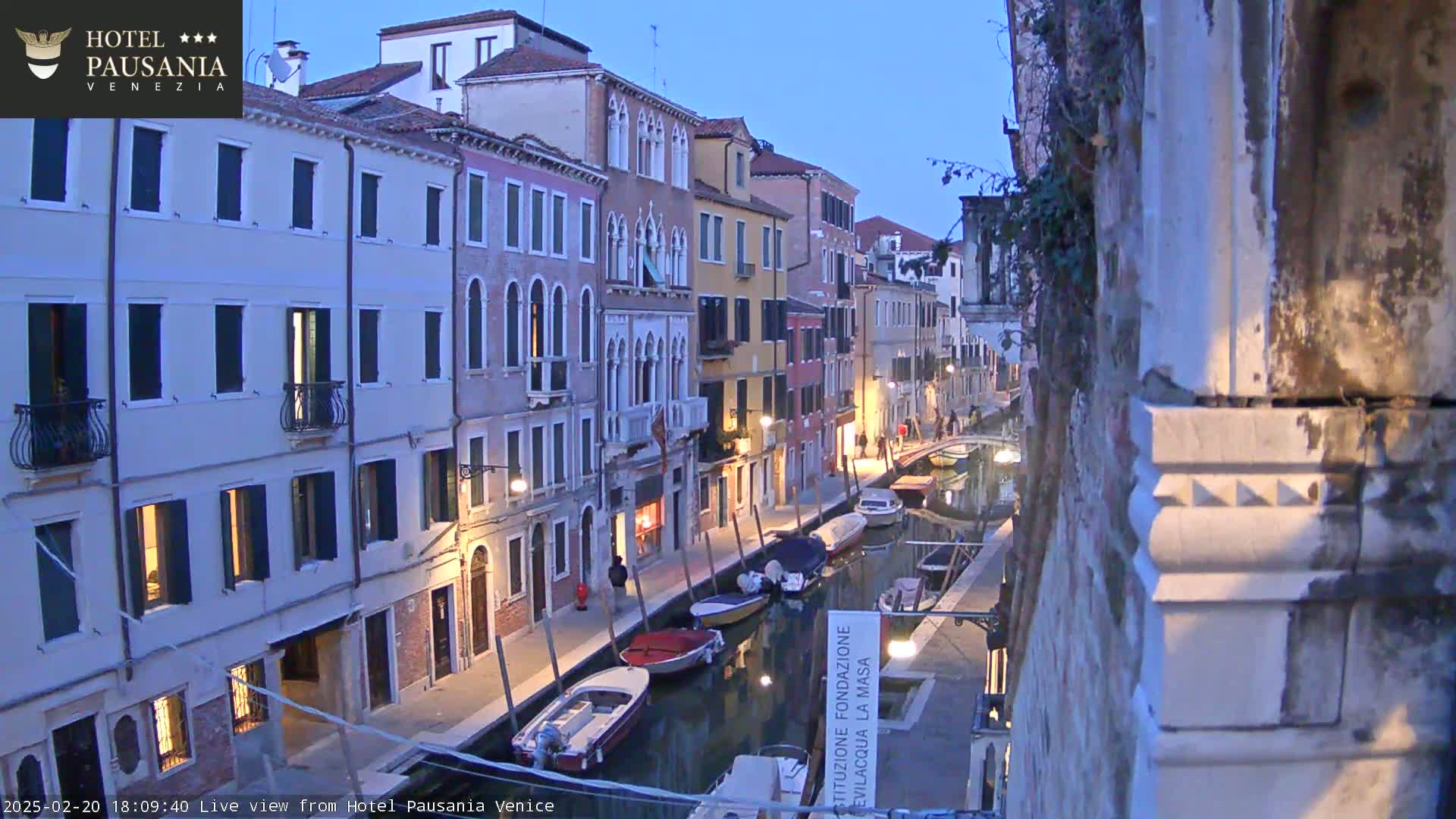 A twilight view down a narrow Venetian canal, lined with closely spaced buildings, showing several moored boats.