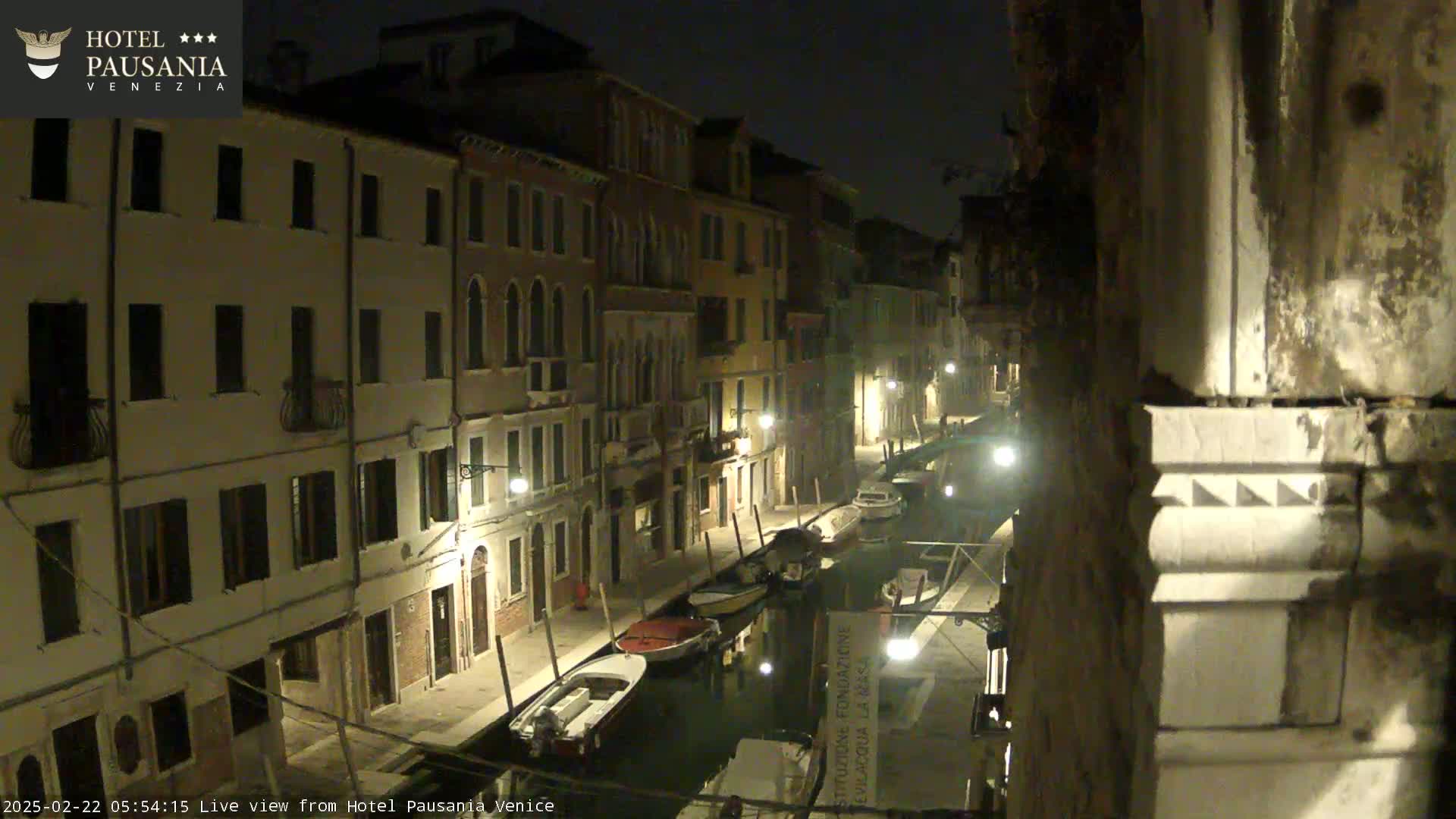 A nighttime view of a narrow canal in Venice, lined with illuminated buildings and several moored boats.