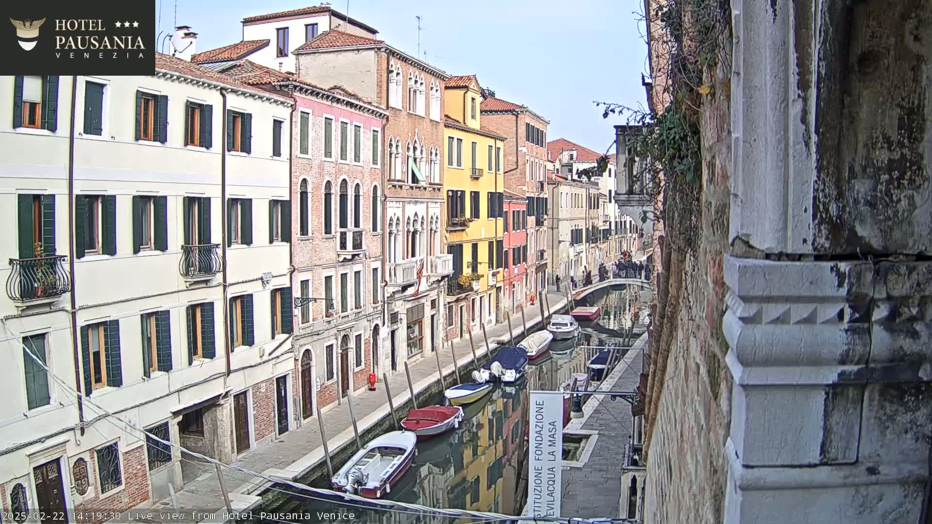 A narrow canal in Venice, lined with colorful buildings, on a partly sunny day with several boats moored along its banks.