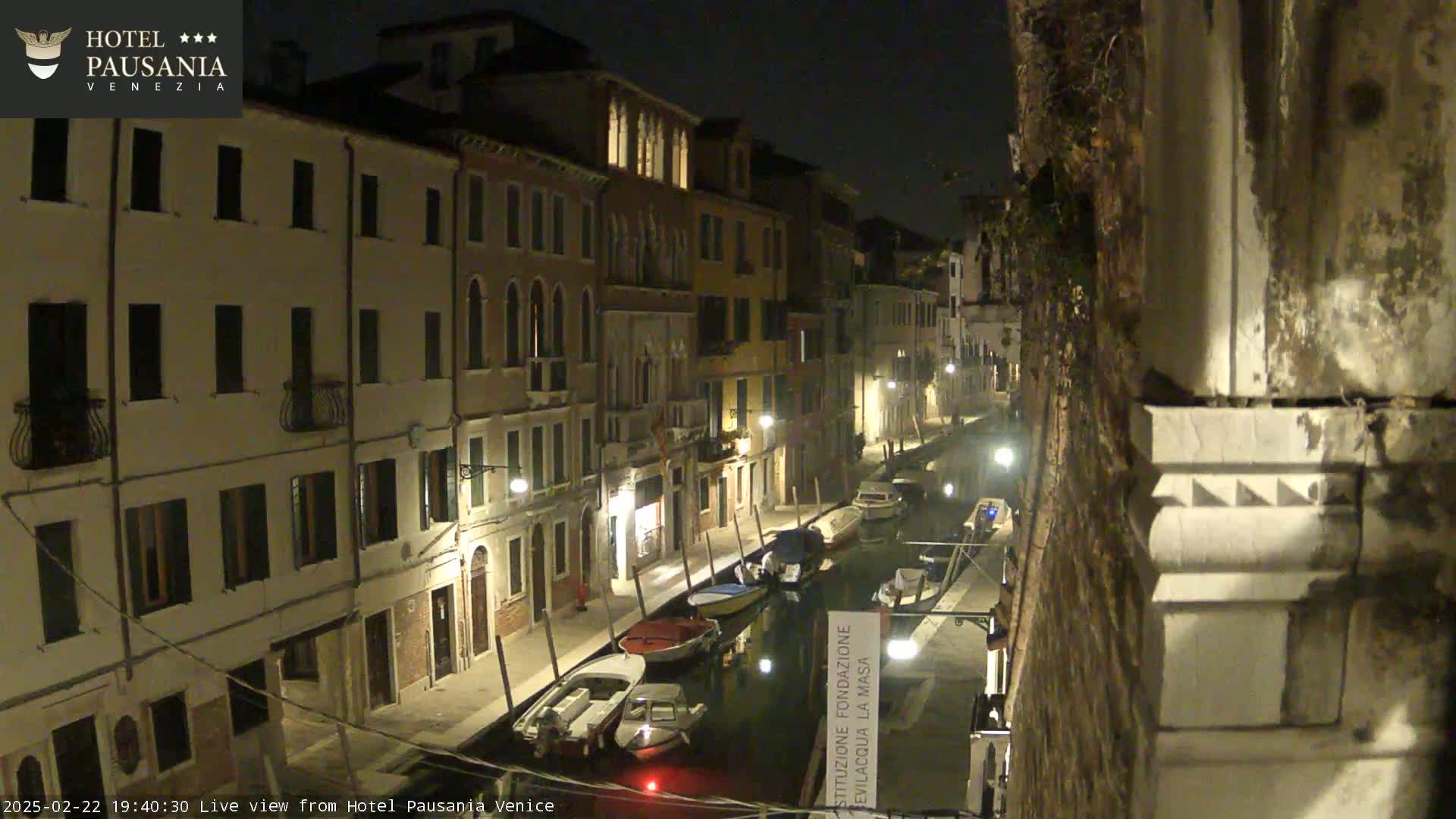 A nighttime view of a canal in Venice, with several boats moored along the sides, flanked by illuminated buildings.