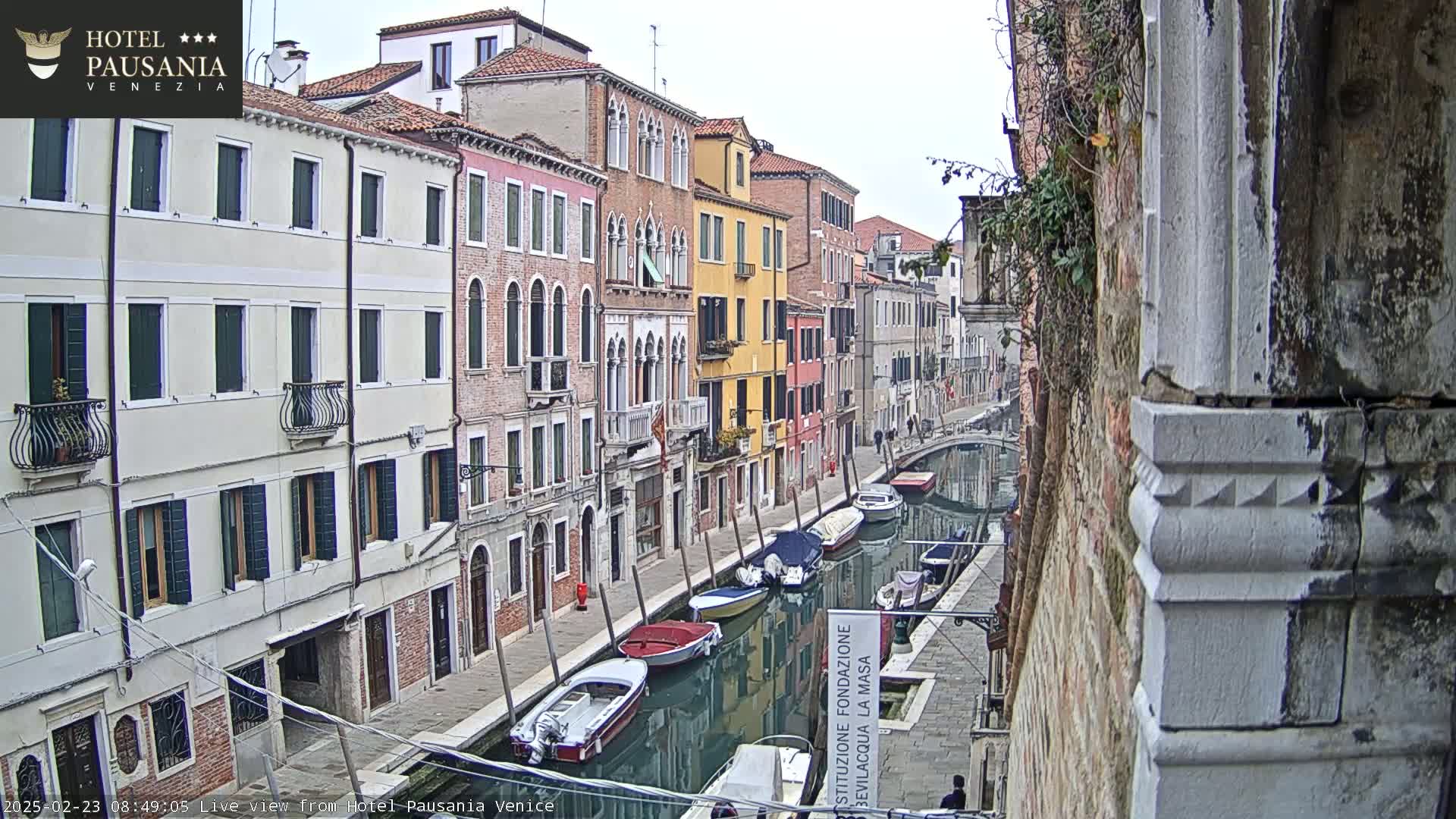 A narrow canal in Venice, lined with closely-spaced buildings of varying colors, shows several boats moored along its calm, reflective waters under an overcast sky.