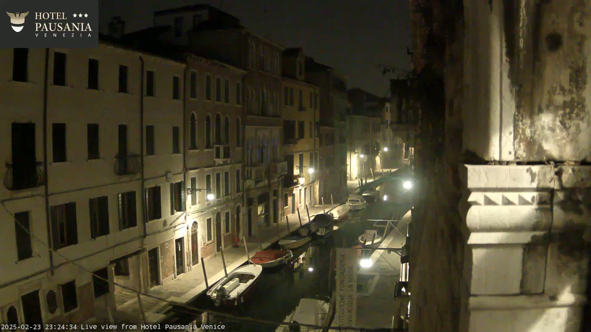 A nighttime view of a narrow canal in Venice, lined with buildings, with several boats moored along the waterway.