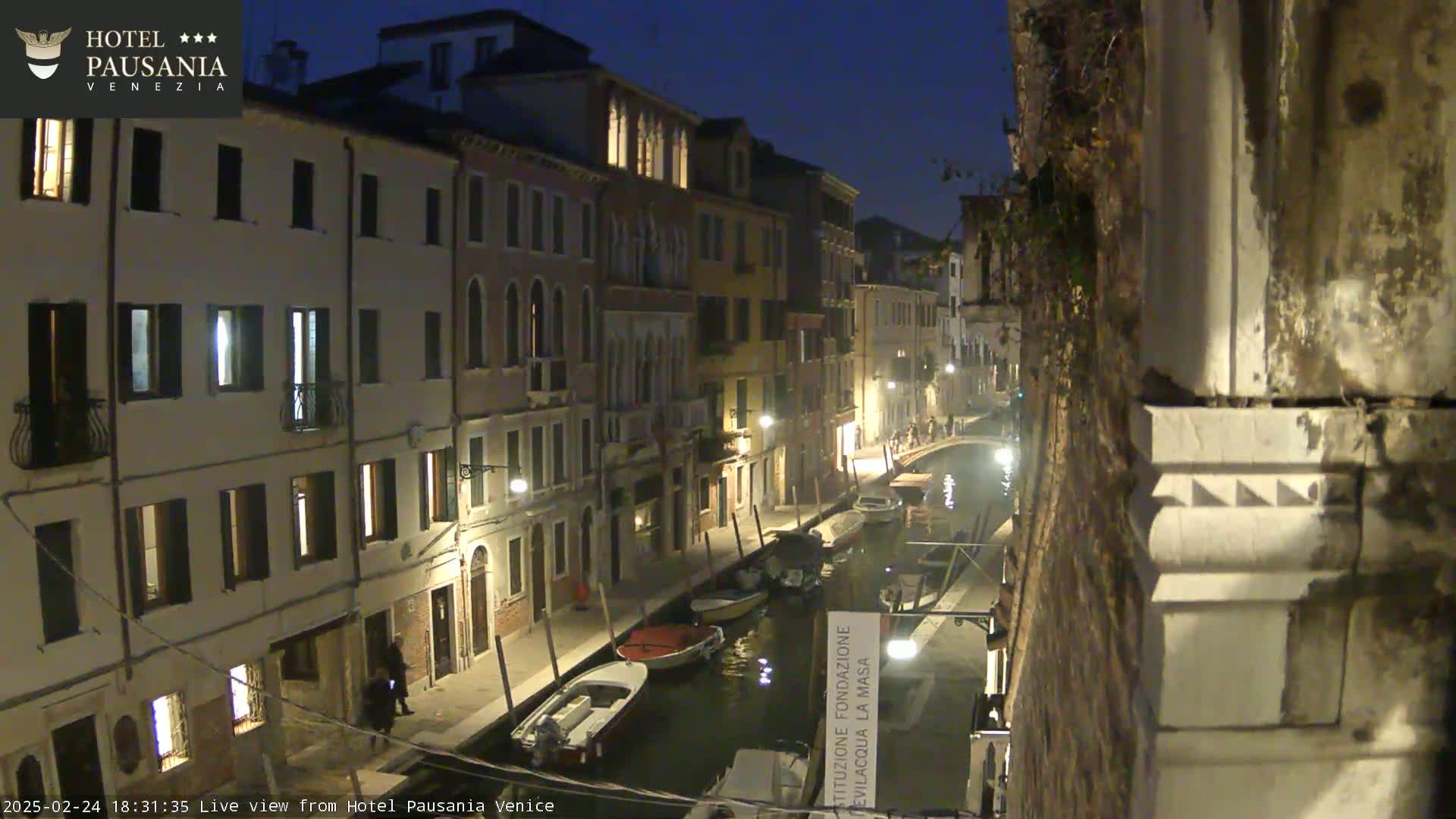 A nighttime view of a narrow canal in Venice, Italy, lined with illuminated buildings, with several small boats moored along the waterway.