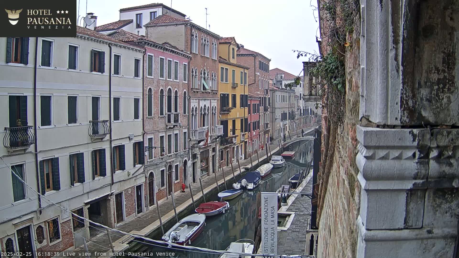 Several boats are moored along a narrow canal in Venice, with colorful buildings lining the waterway under an overcast sky.