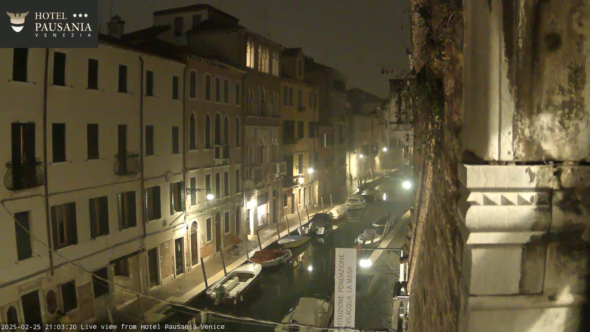 A nighttime view of a canal in Venice, with several boats moored along its banks, under foggy or misty conditions.