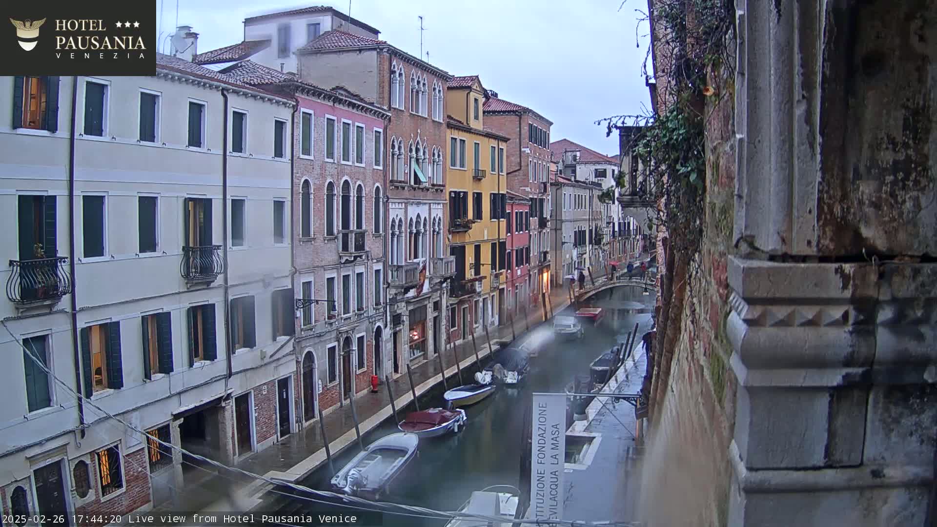 A rain-slicked canal in Venice, Italy, is lined with tall, narrow buildings, and several motorboats are moored along its edge.