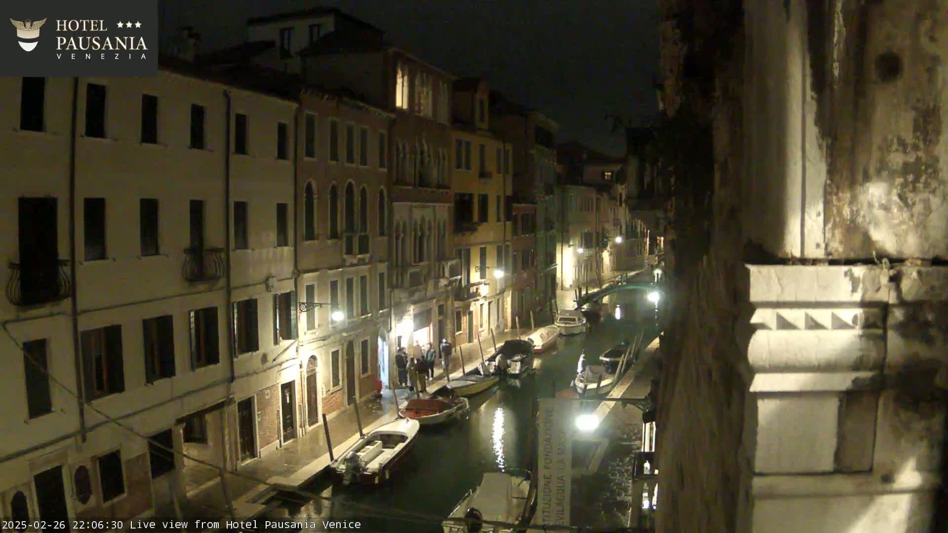 A nighttime view of a narrow canal in Venice, with several boats moored along the sides and buildings lit along the waterway.