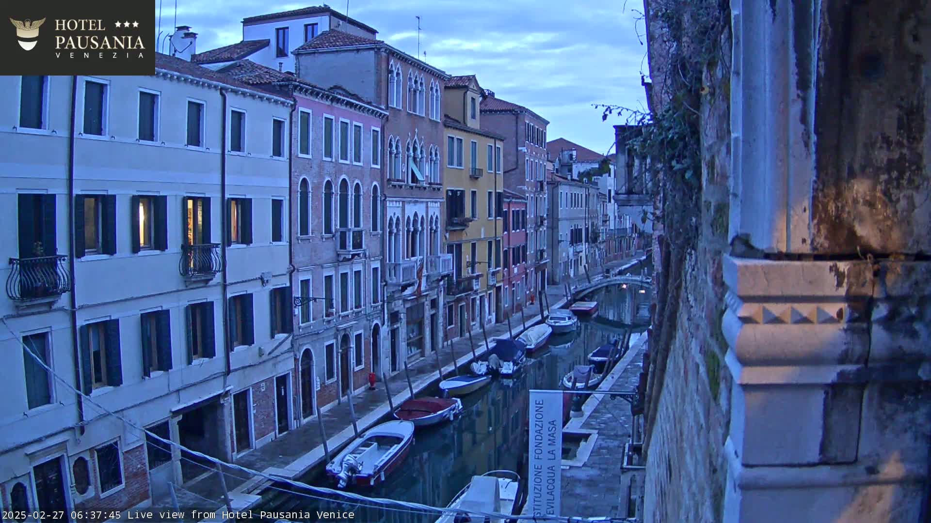 A narrow canal in Venice, Italy, is lined with closely spaced buildings at dawn under a twilight sky, with several small boats moored along the waterway.