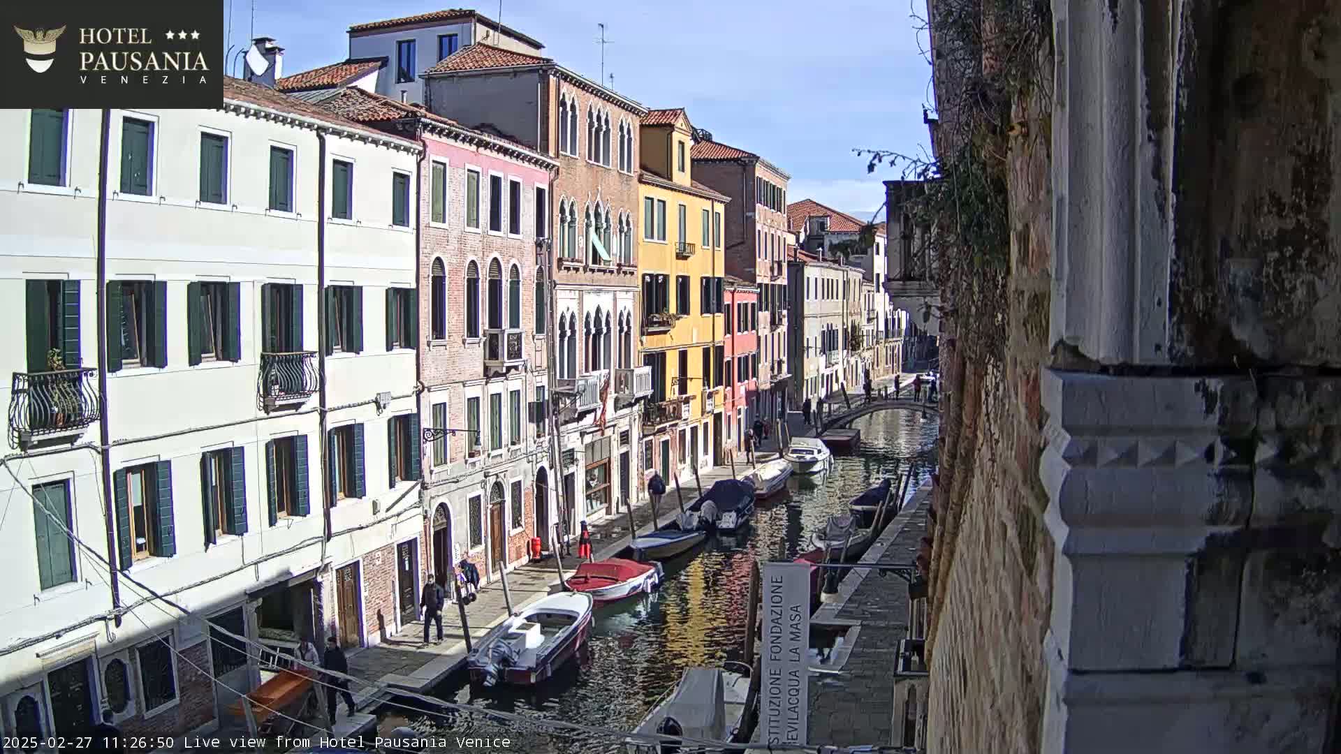 A sunny view down a canal in Venice, showing colorful buildings and several boats moored along the waterway.