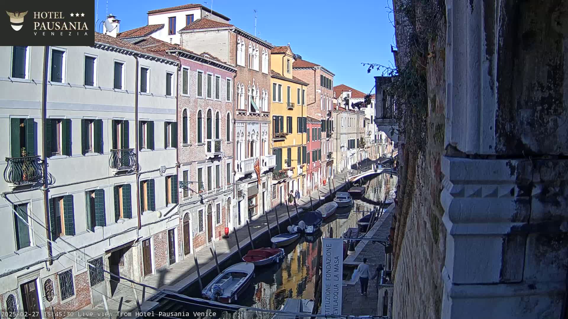 A sunny day in Venice shows a narrow canal lined with colorful buildings, several boats moored along the waterway, and a few pedestrians walking along the canal-side path.